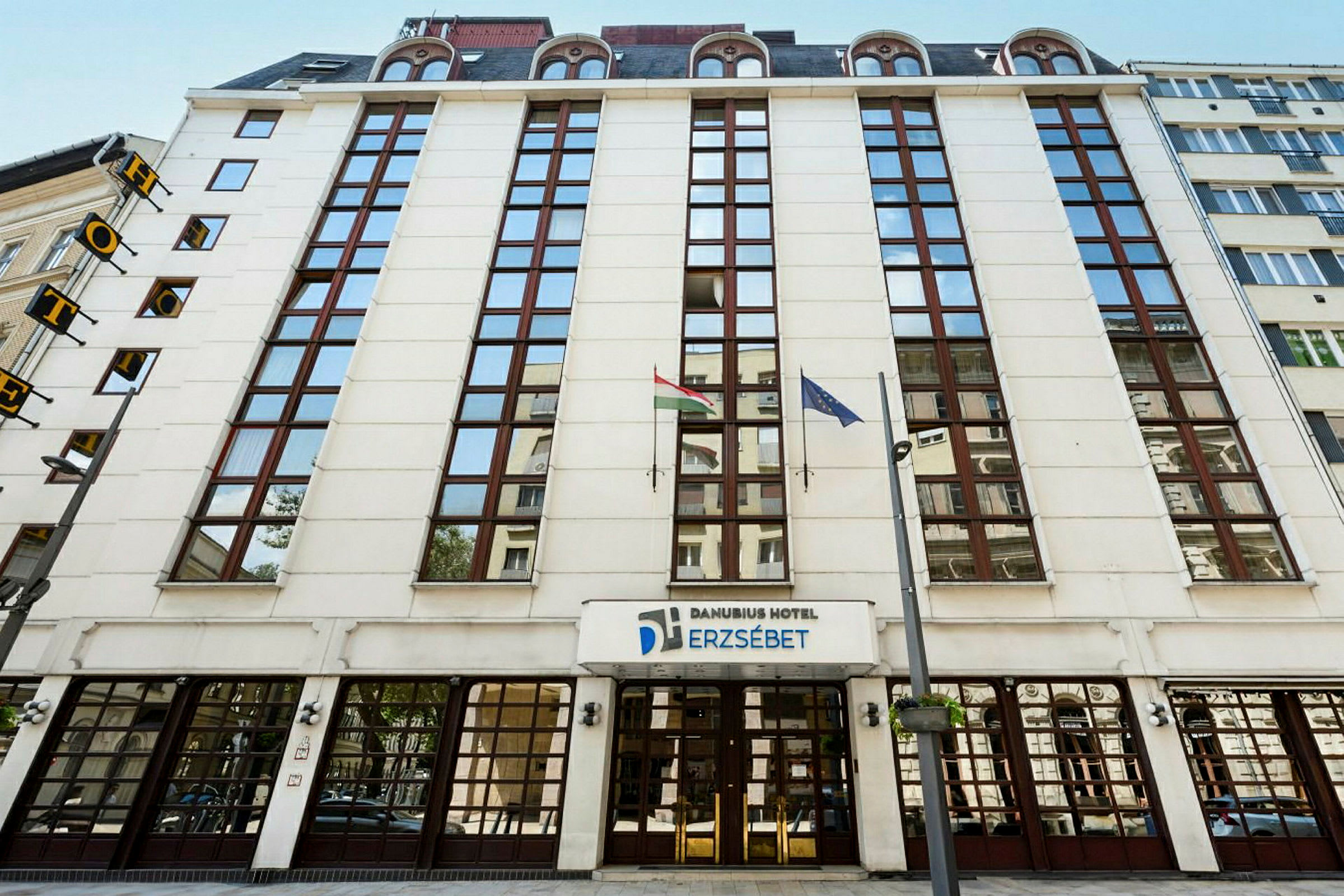 Forsiden av Danubius Hotel Erzsébet med glassvinduer og flagg. (AI-generert tekst)