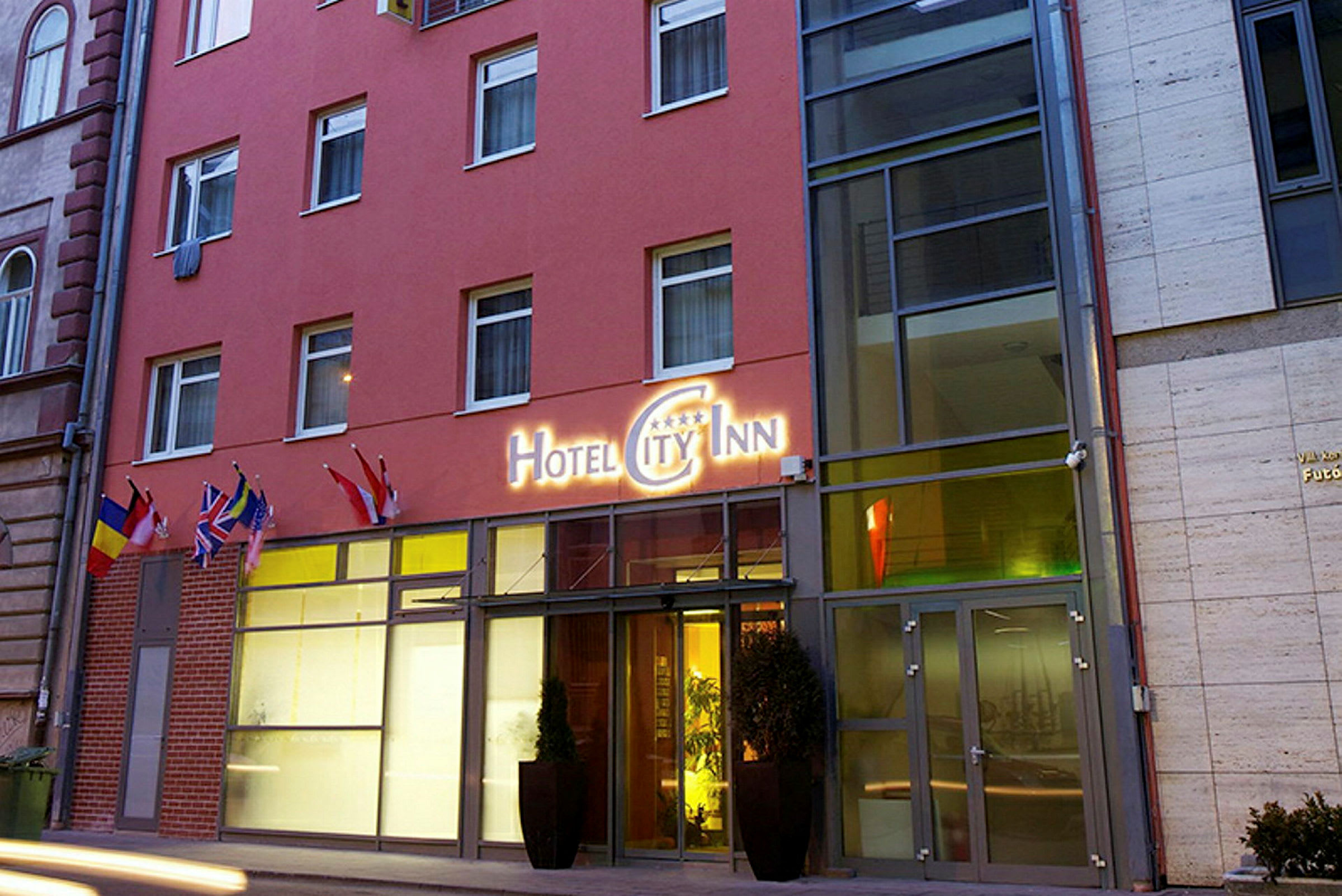 Utenfor en hotellbygning med et skilt som lyder "Hotel City Inn" og flere flagg ved inngangen. (AI-generert tekst)