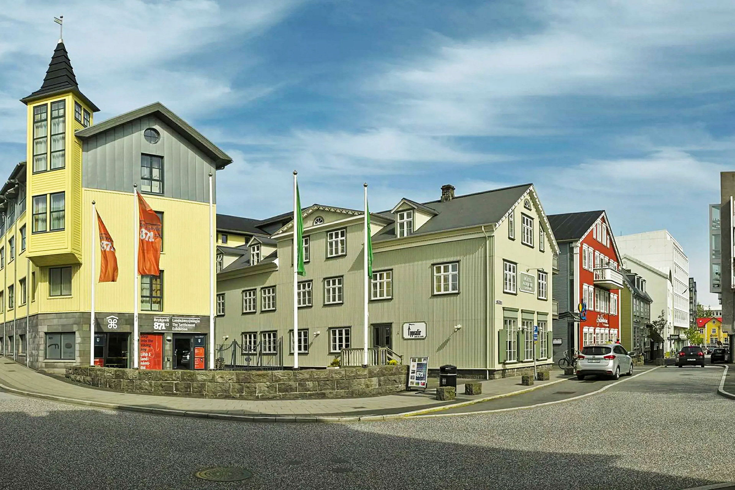 En gate med bygninger i forskjellige farger, inkludert en gul bygning med et tårn og flaggstenger foran. (AI-generert tekst)