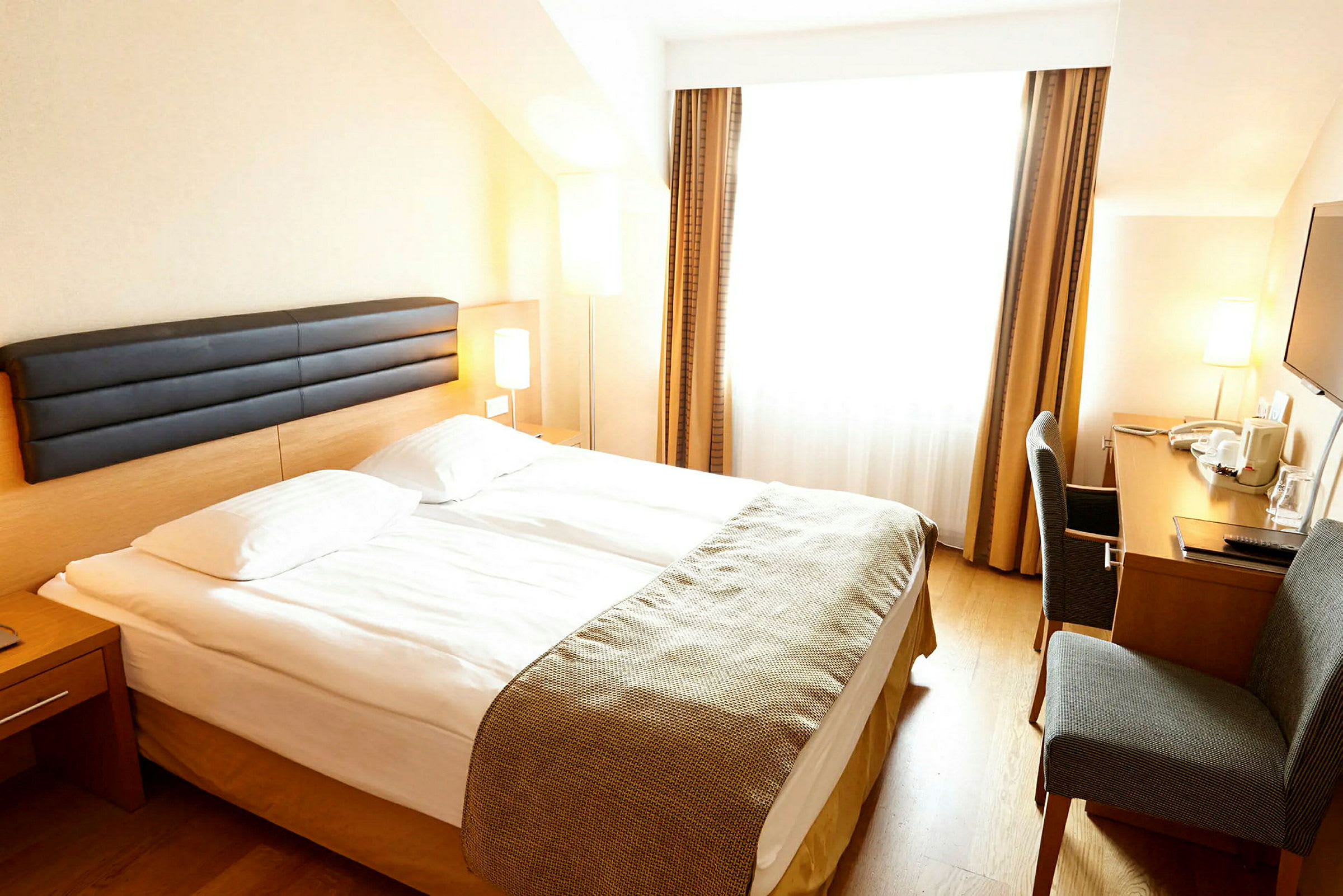 Hotellrom med en dobbeltseng, et skrivebord og en stol. Rommet har store vinduer med gardiner. (AI-generert tekst)