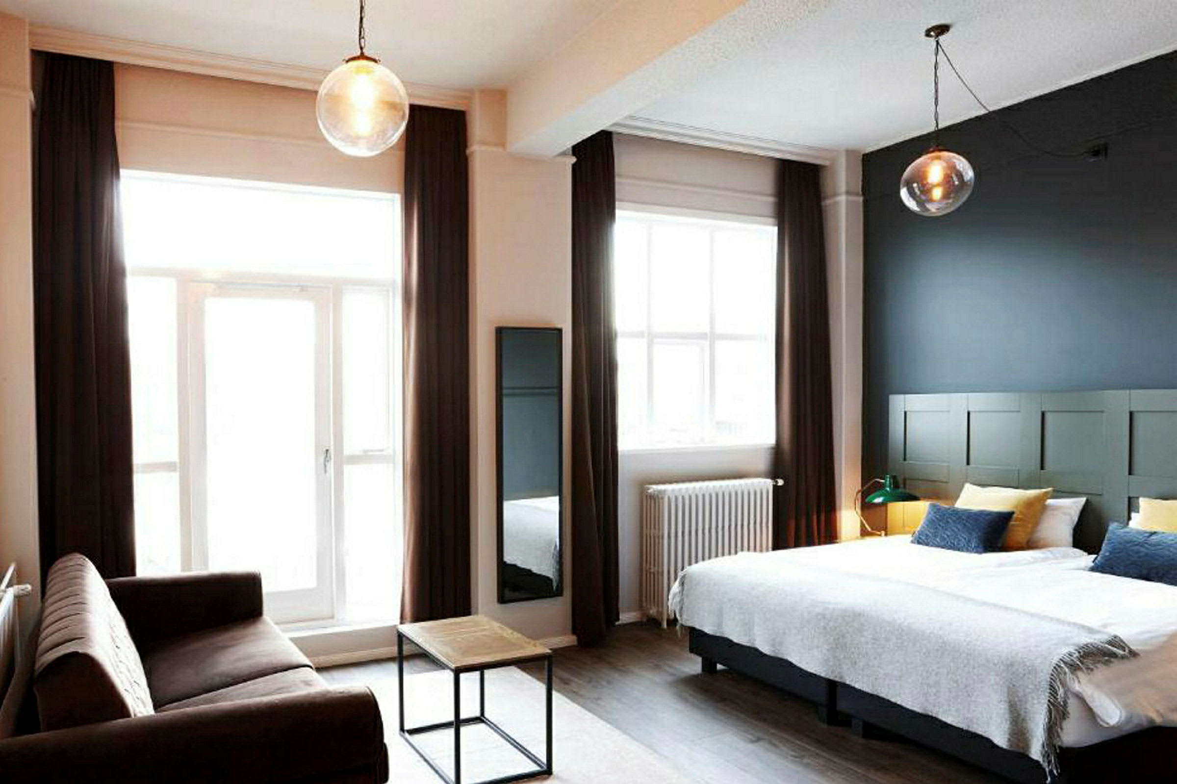 Et hotellrom med en dobbeltseng, en sofa og et lite salongbord samt store vinduer med gardiner. (AI-generert tekst)