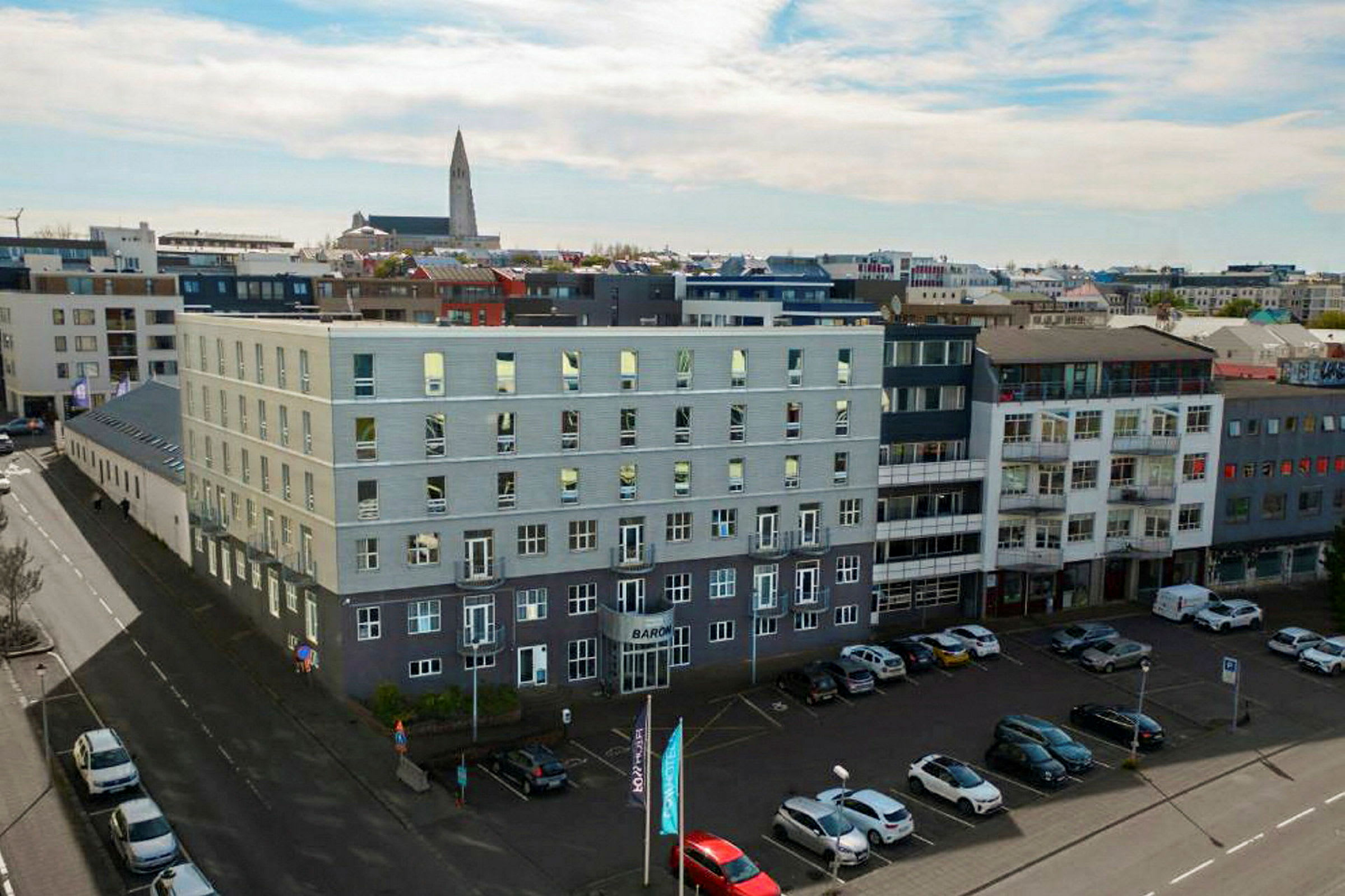 Flere moderne bygninger omgir et åpent parkeringsområde, med biler parkert på bakken og en kirke synlig i bakgrunnen. (AI-generert tekst)