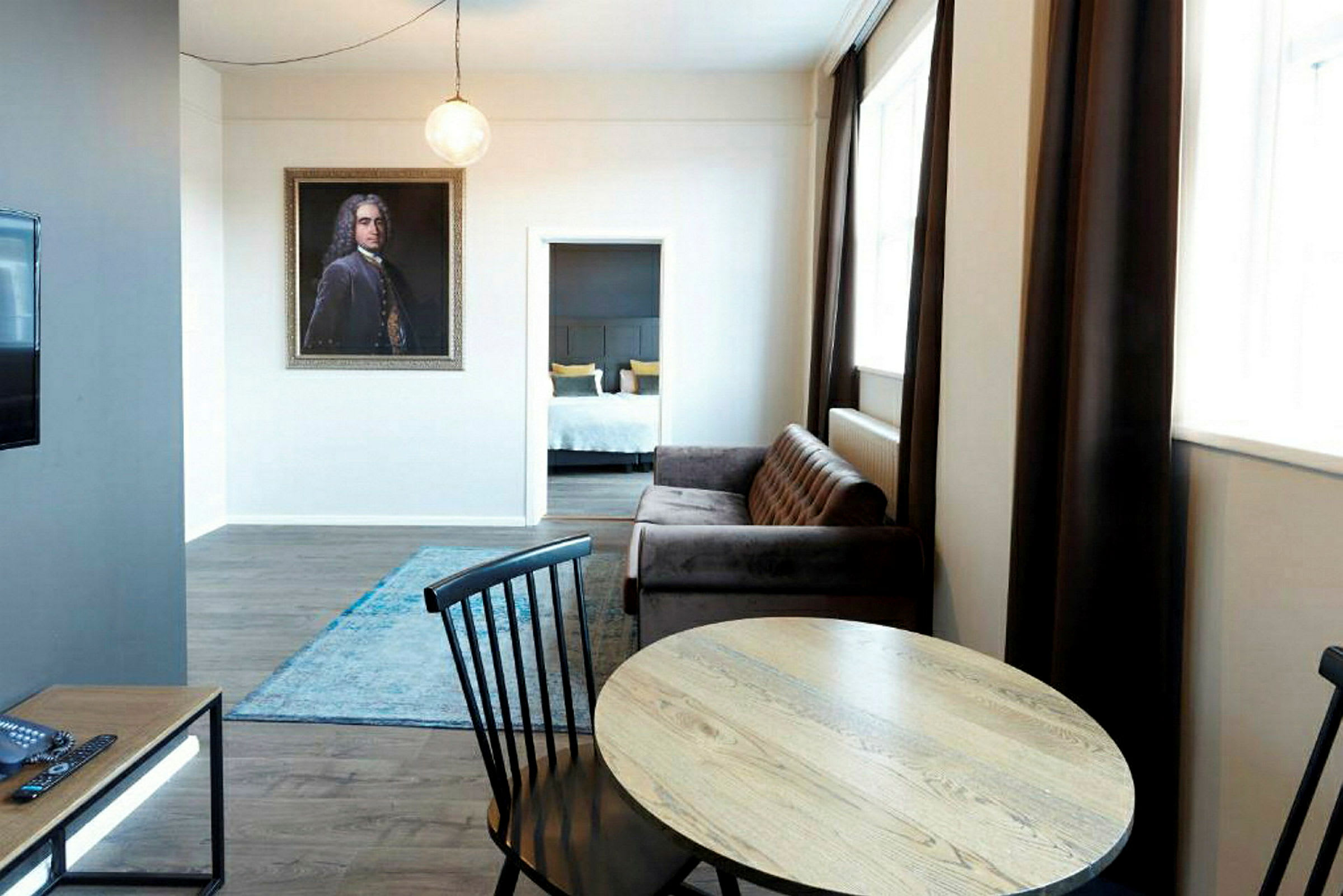 En stue med bord og stoler foran en sofa, et teppe på gulvet og en åpning til et soverom i bakgrunnen. (AI-generert tekst)