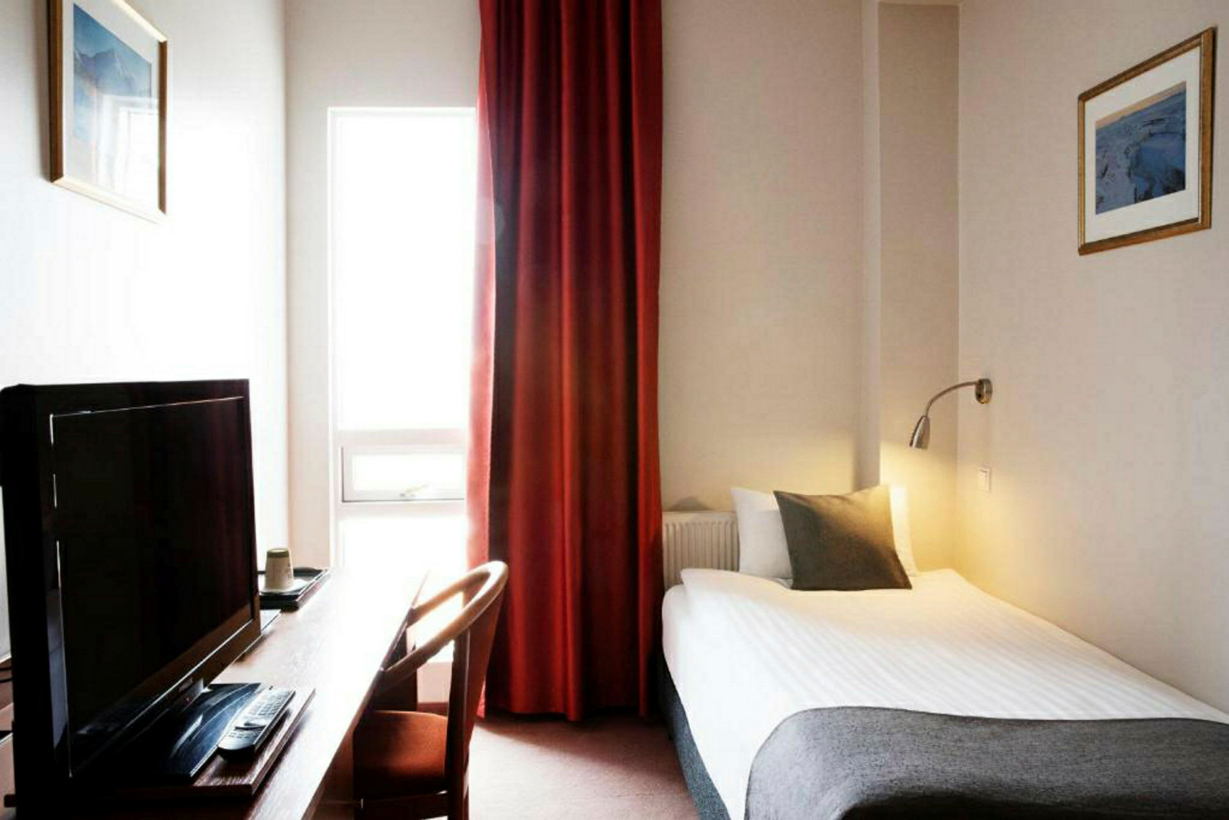 Et hotellrom med en enkeltseng, skrivebord, TV og vindu med røde gardiner. (AI-generert tekst)