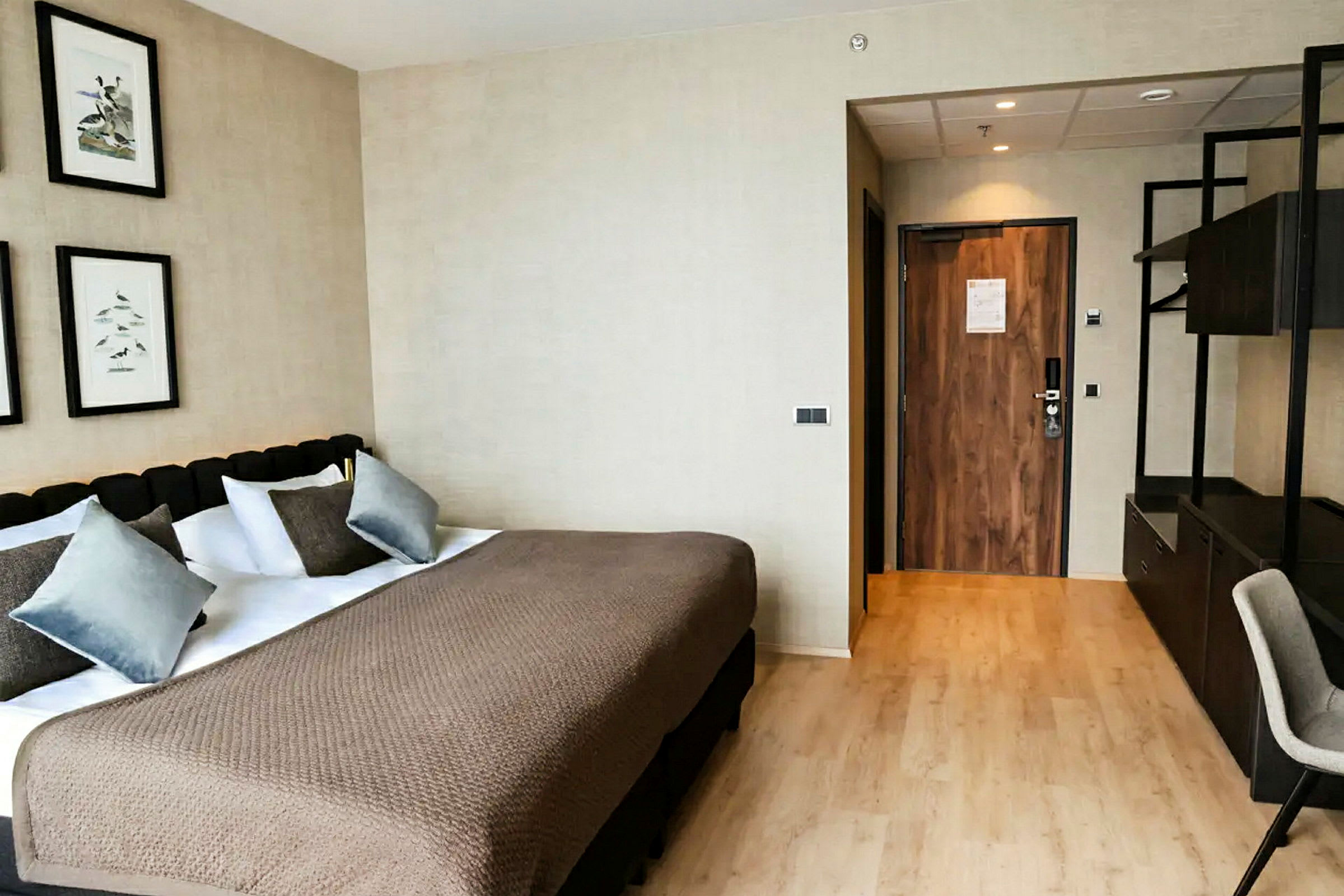 Et hotellrom med en stor seng, flere puter og et beige sengeteppe. På veggen ovenfor henger bilder, og en dør til høyre leder ut. (AI-generert tekst)
