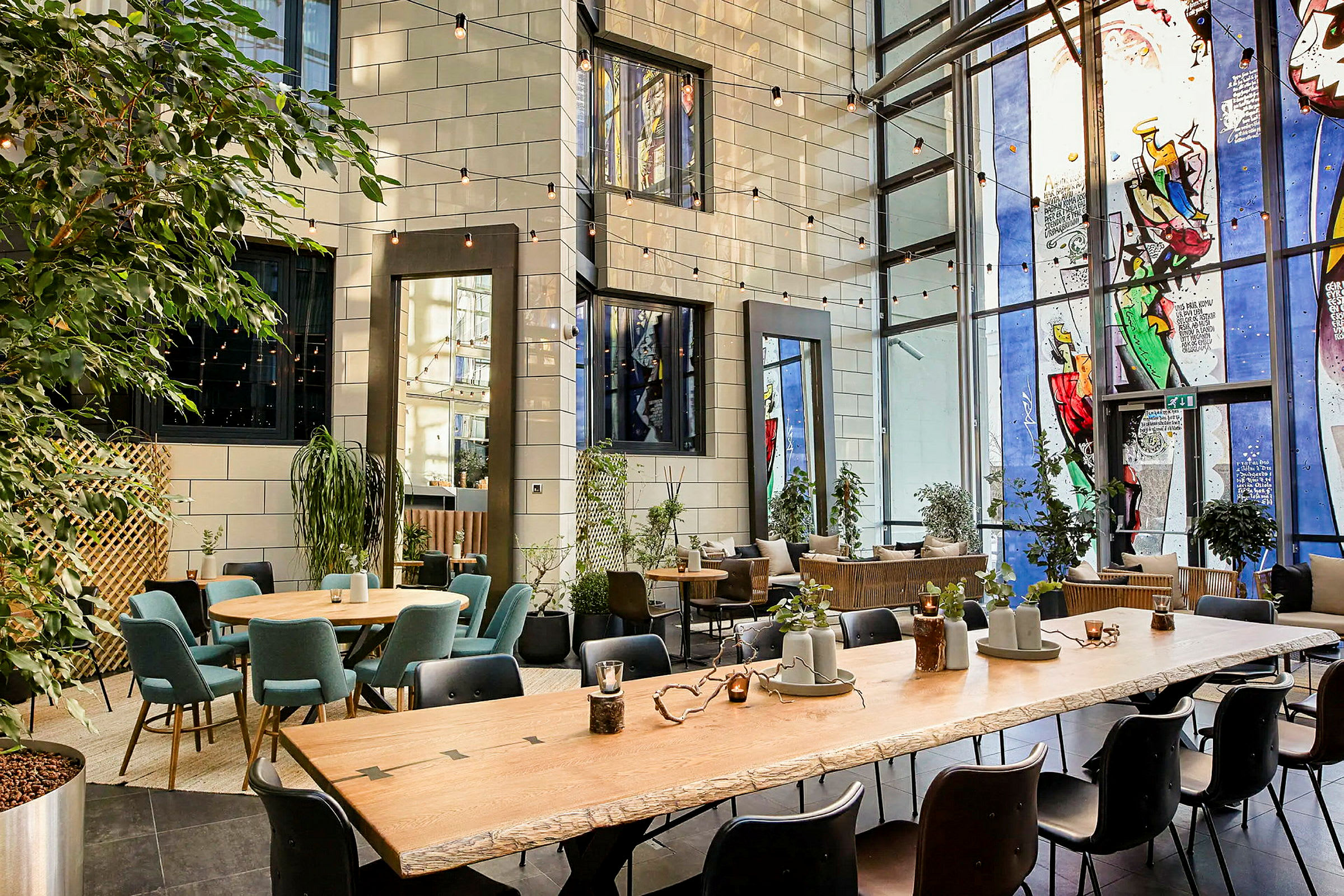 En innendørs lounge med store vinduer, sitteplasser rundt flere bord og grønne planter plassert i ulike områder. (AI-generert tekst)