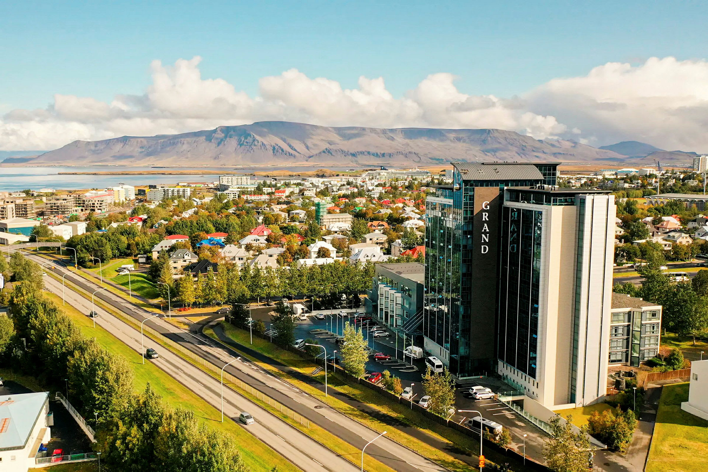 Et bymiljø med en motorvei i forgrunnen, trær, bygninger og et større hotell. I bakgrunnen sees et fjellandskap. (AI-generert tekst)