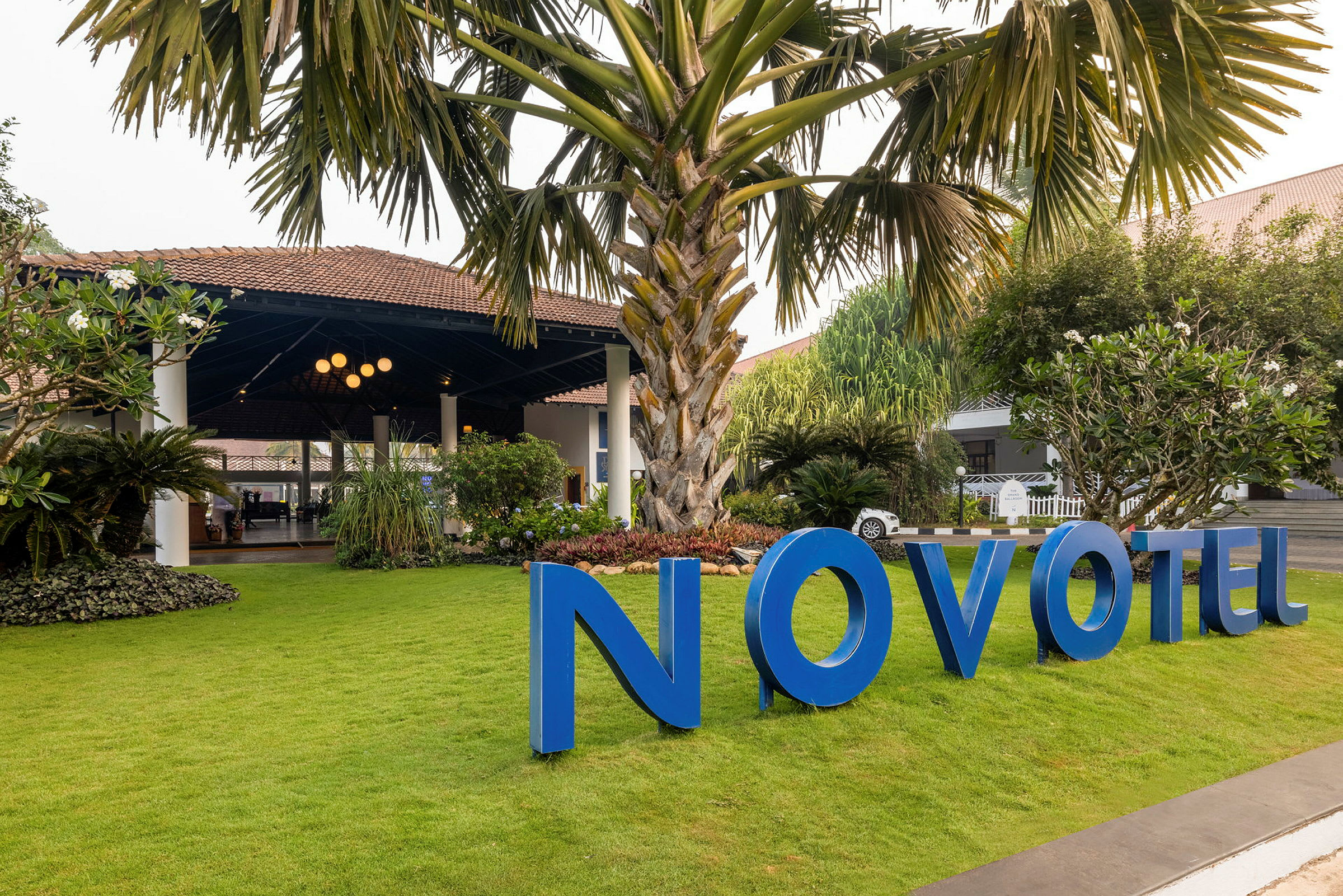 En hage med en stor palme og grønt gress foran en bygning med tak og blått skilt som sier "Novotel". (AI-generert tekst)