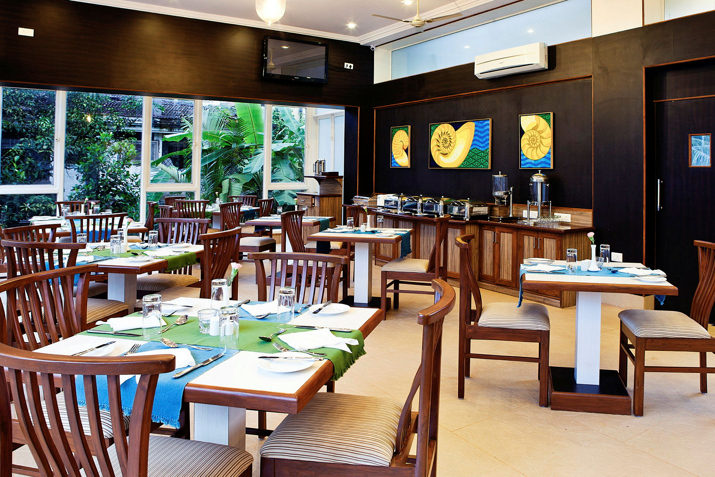 En restaurant med trebord og stoler, fargerike bordduk dekorasjoner. Bakgrunnen viser en buffet med serveringsbeholdere. Vinduer gir utsikt mot grønne planter. (AI-generert tekst)
