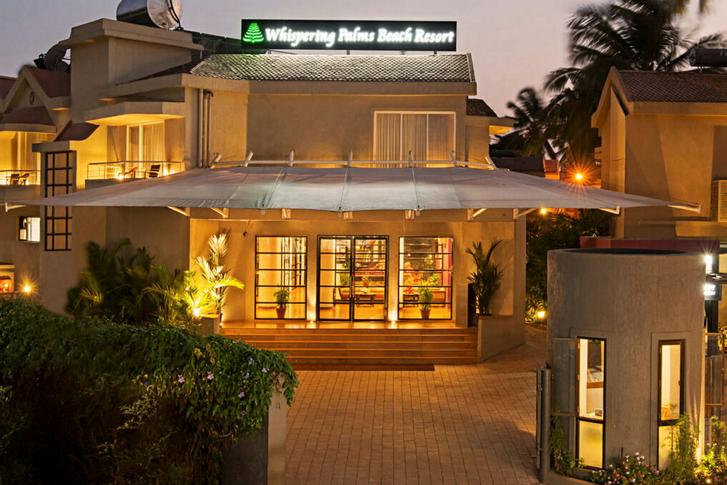Inngangen til Whispering Palms Beach Resort med en opplyst fasade og grønne planter. (AI-generert tekst)