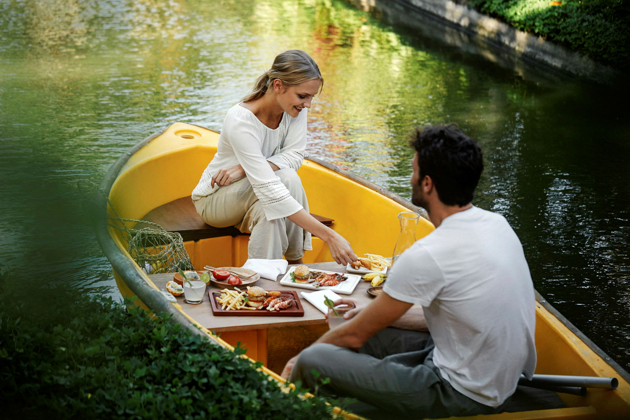 To personer sitter i en gul robåt på en stille kanal og spiser et måltid ved et bord. (AI-generert tekst)
