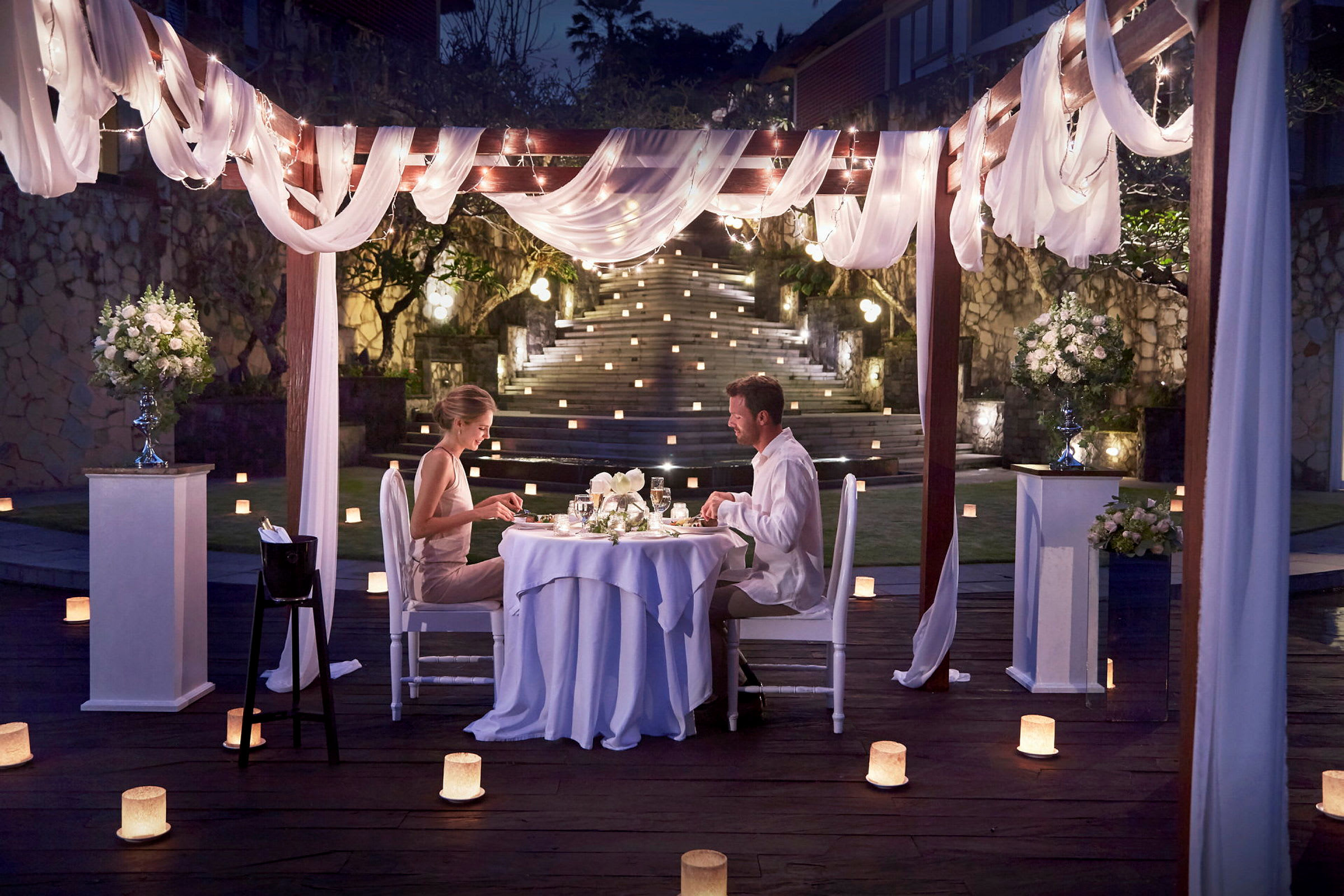 Et utendørs middagsbord under en elegant dekorert pergola med lys og blomster. (AI-generert tekst)