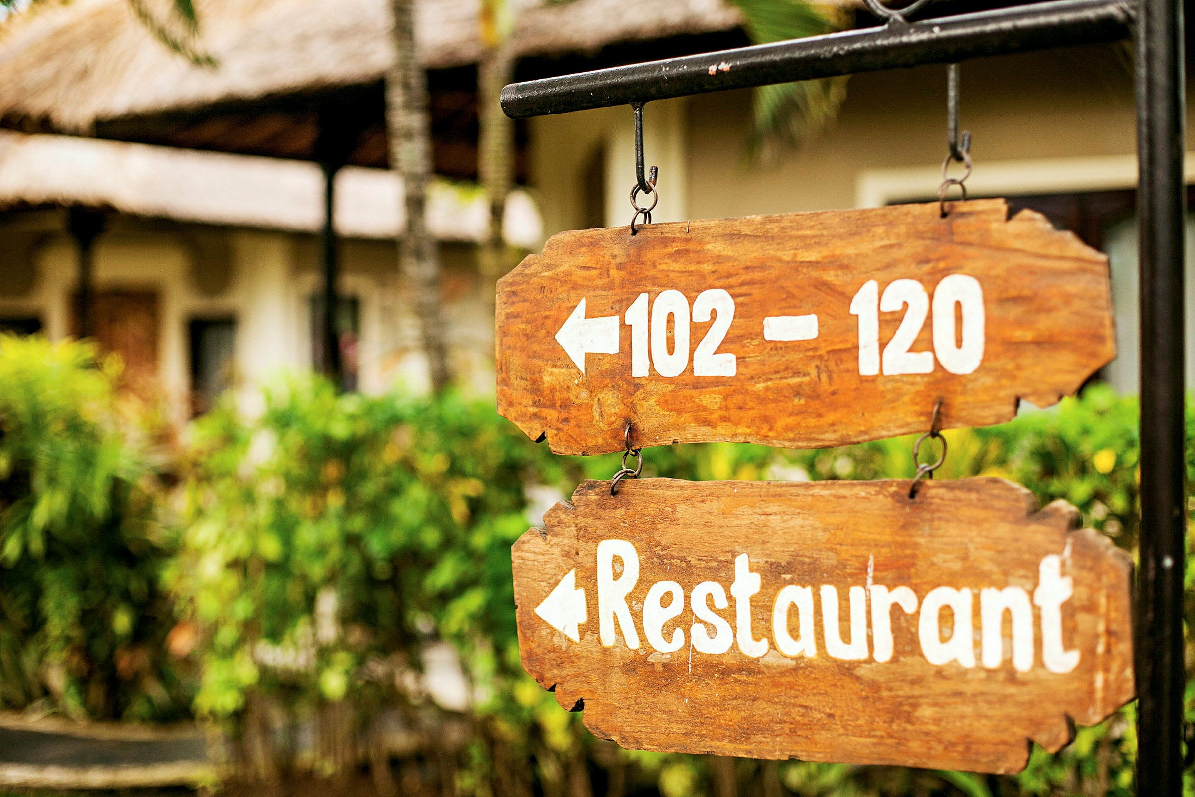 To treskilt med hvit tekst peker til venstre, viser tallene 102–120 og ordet restaurant. Bakgrunnen har grønne planter og bygninger. (AI-generert tekst)