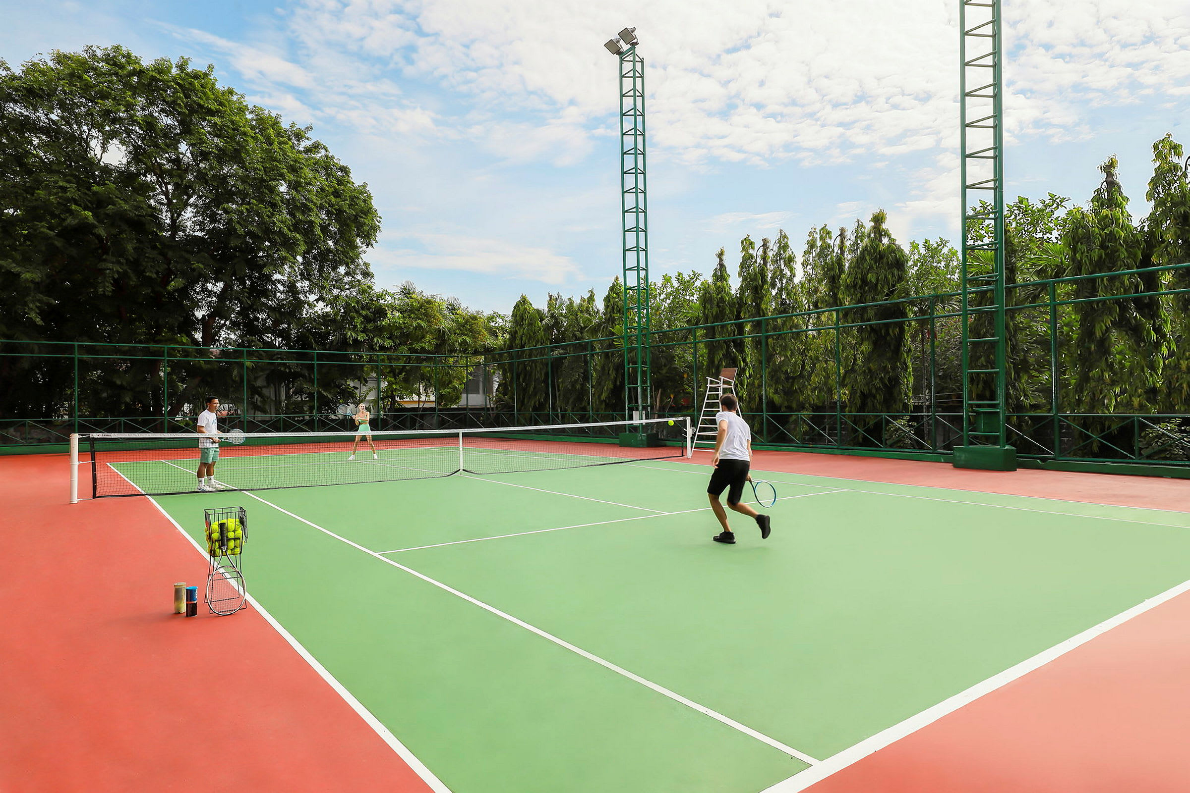 Personer spiller tennis på en utendørs bane. I bakgrunnen sees trær. (AI-generert tekst)