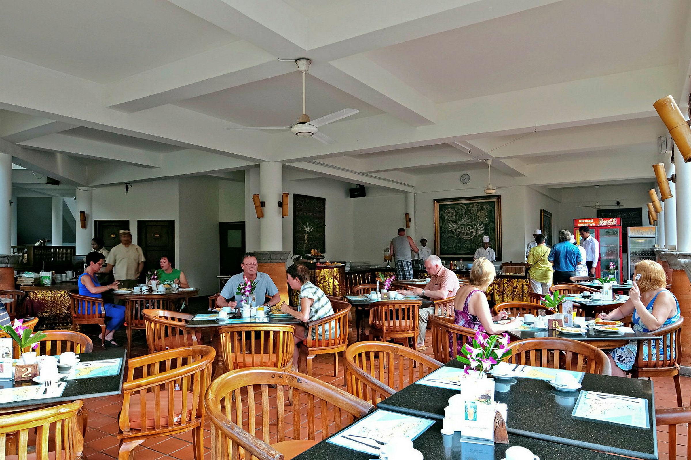 Et restaurantområde med bord og stoler, hvor folk sitter og spiser. Innredningen er lys med hvite vegger og tak. (AI-generert tekst)