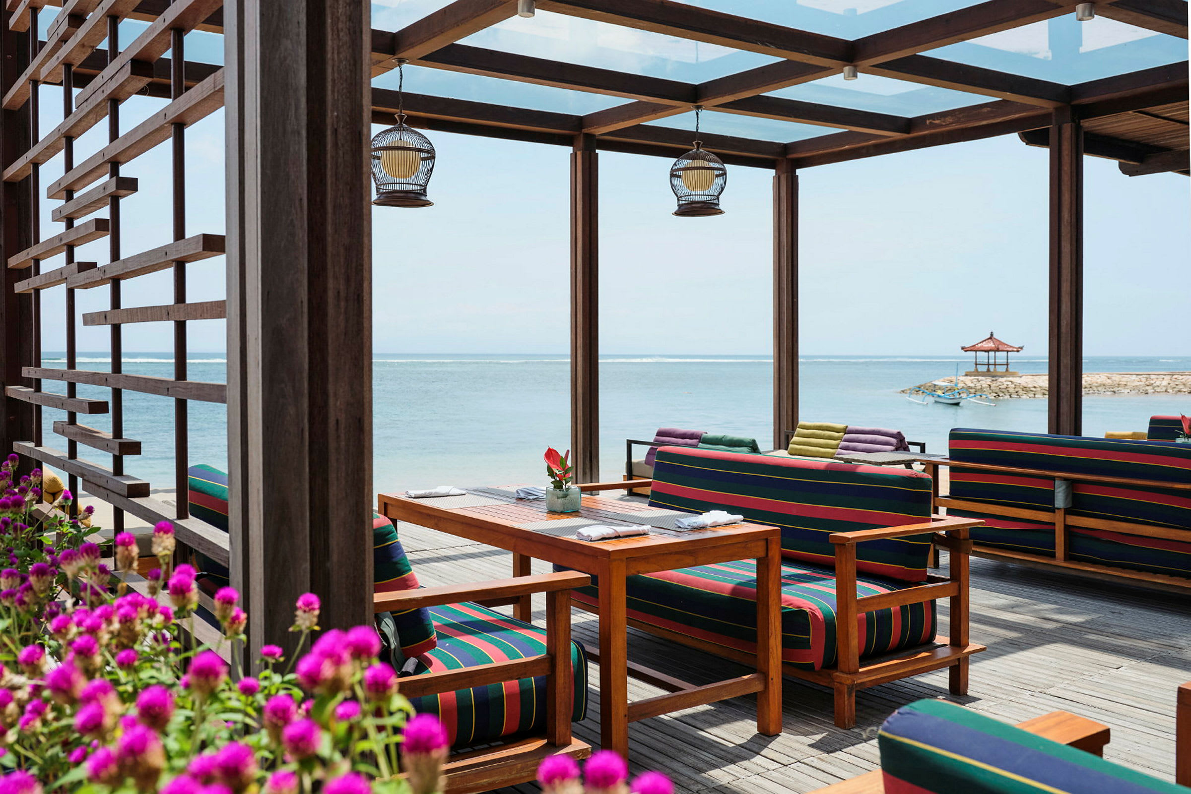Utendørsområde med møbler med fargerike striper, blomster i forgrunnen og sjøutsikt med strandpaviljong. (AI-generert tekst)