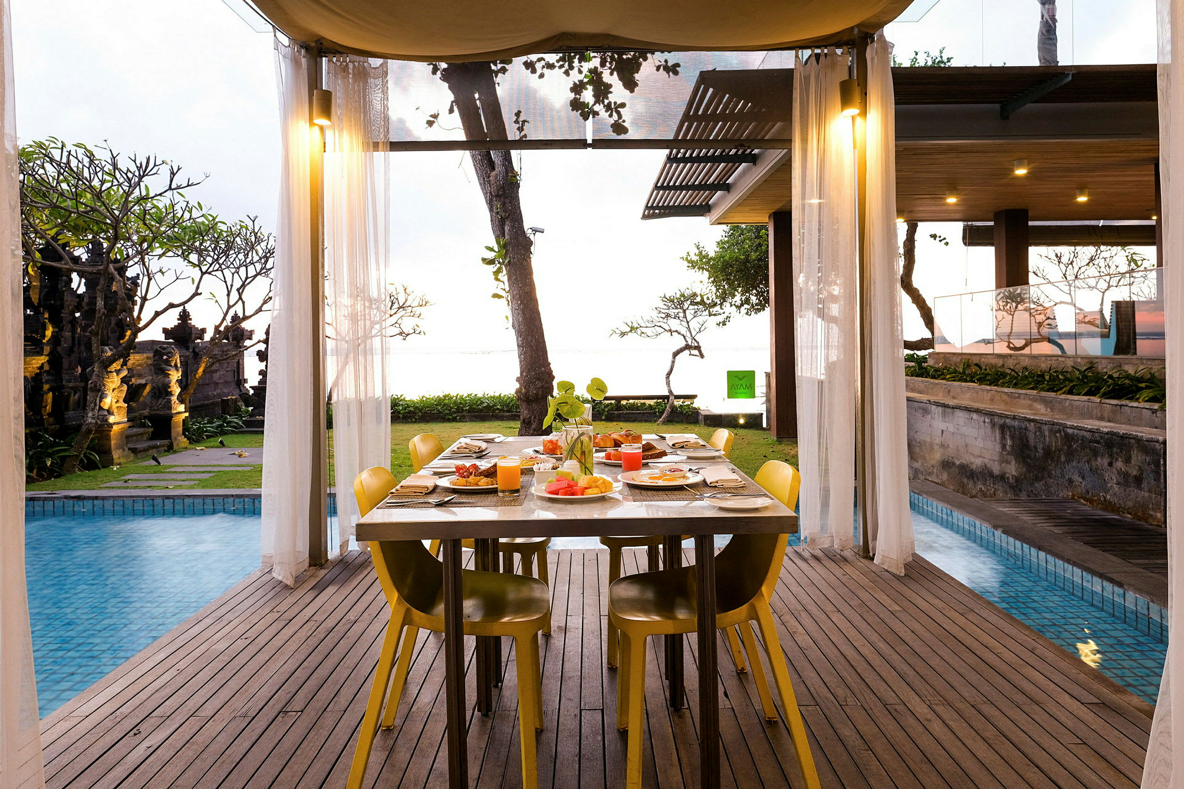 Et bord dekket til måltid står under en overbygd terrasse nær et basseng med utsikt mot hagen og havet. (AI-generert tekst)