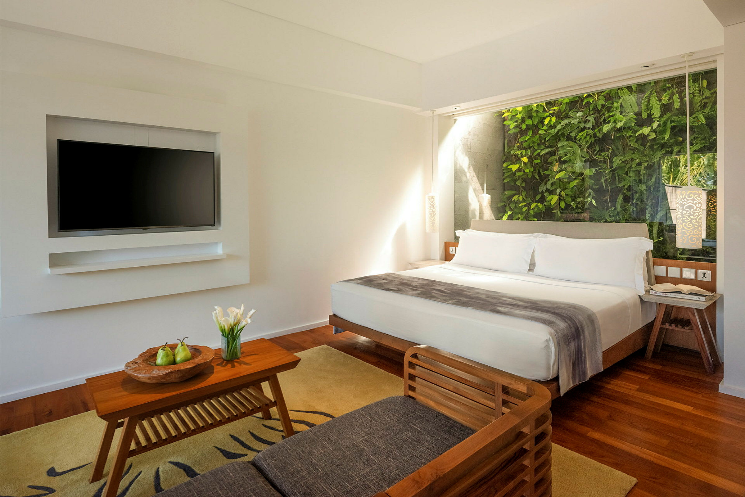 Et moderne hotellrom med en stor seng, en sofa og et salongbord foran en veggmontert TV. Bak sengen er et vindu med grønne planter. (AI-generert tekst)