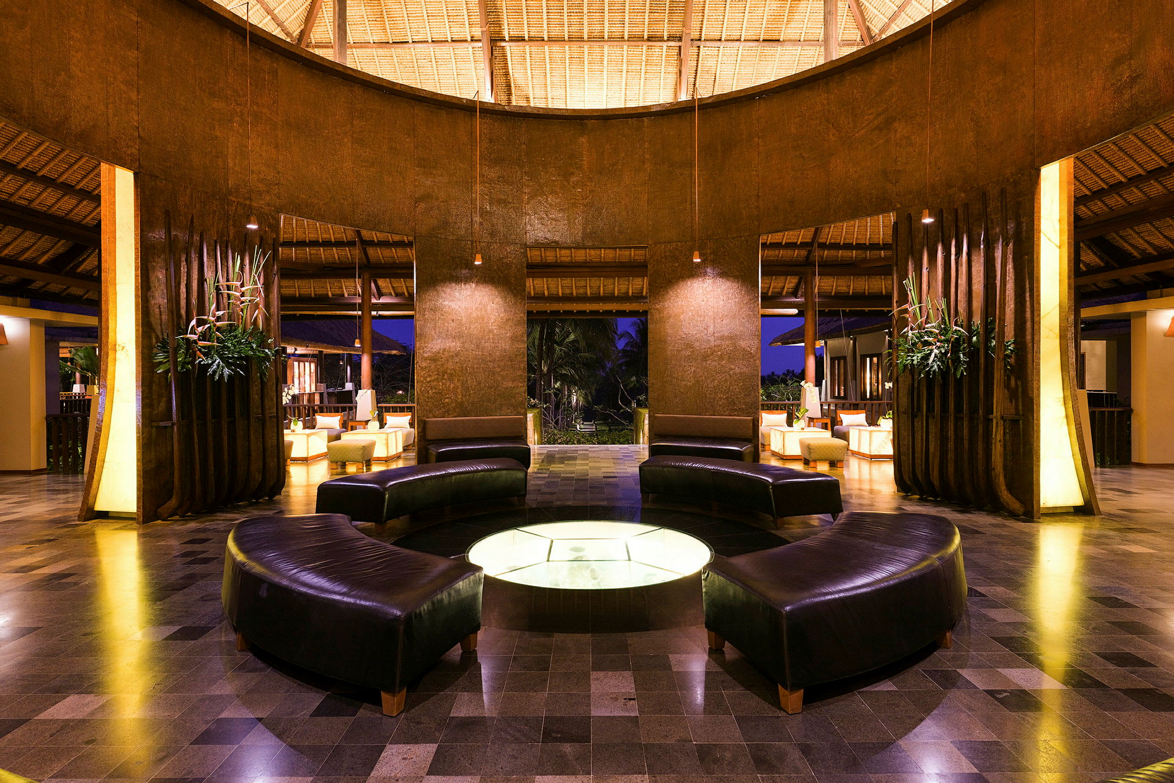 En elegant lobby med mørke sofaer arrangert i en sirkel rundt et opplyst bord, omgitt av trepanel og planter. (AI-generert tekst)