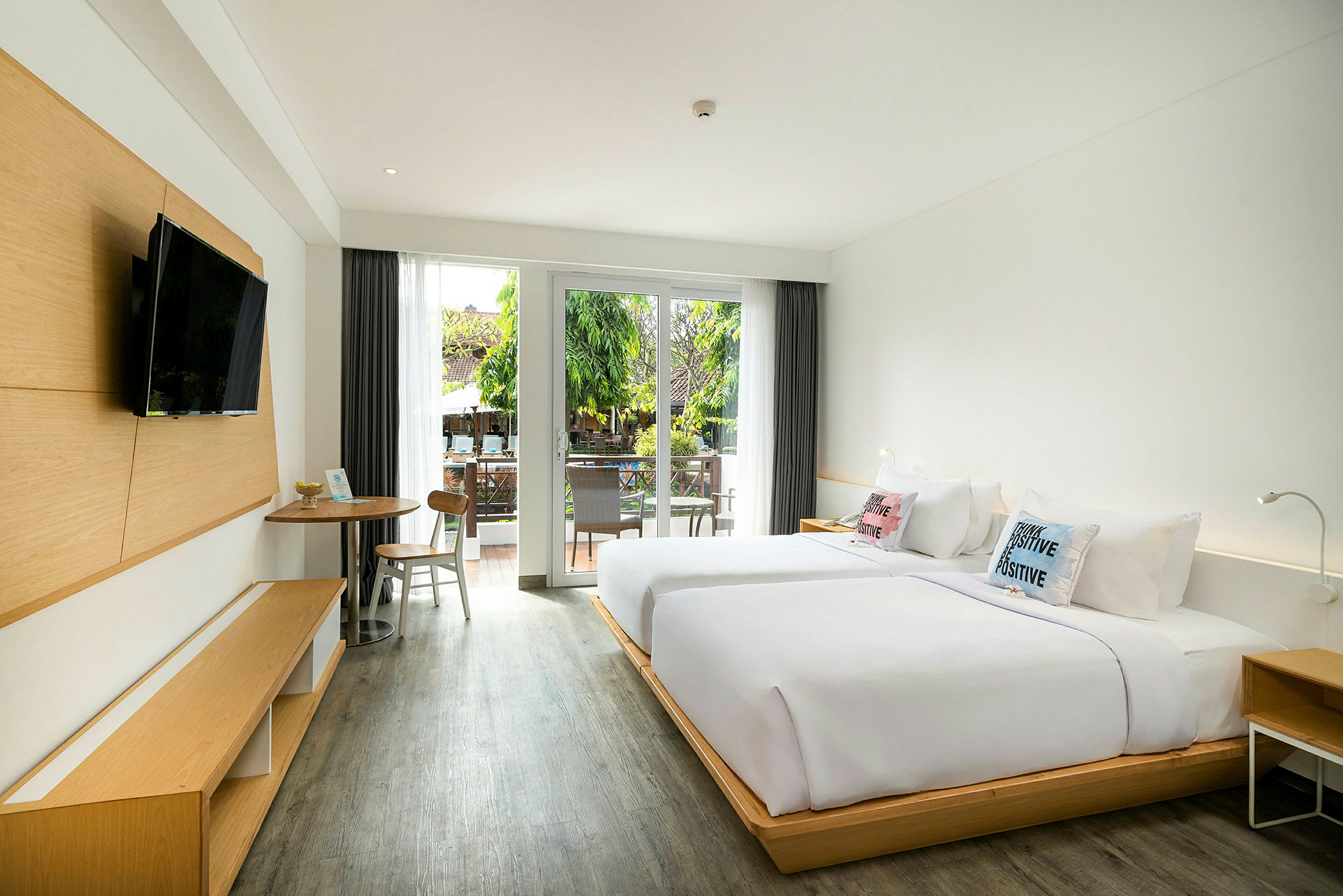 Et hotellrom med to senger, et bord med stoler og en moderne TV på veggen. Rommet har store glassdører som fører ut til en balkong med utsikt over grønne omgivelser. (AI-generert tekst)