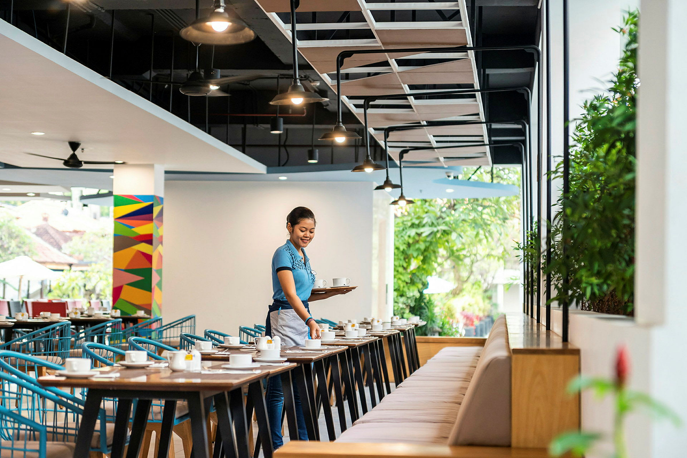 Et restaurantlokale med bord og stoler, der en person bærer et brett med kopper. (AI-generert tekst)