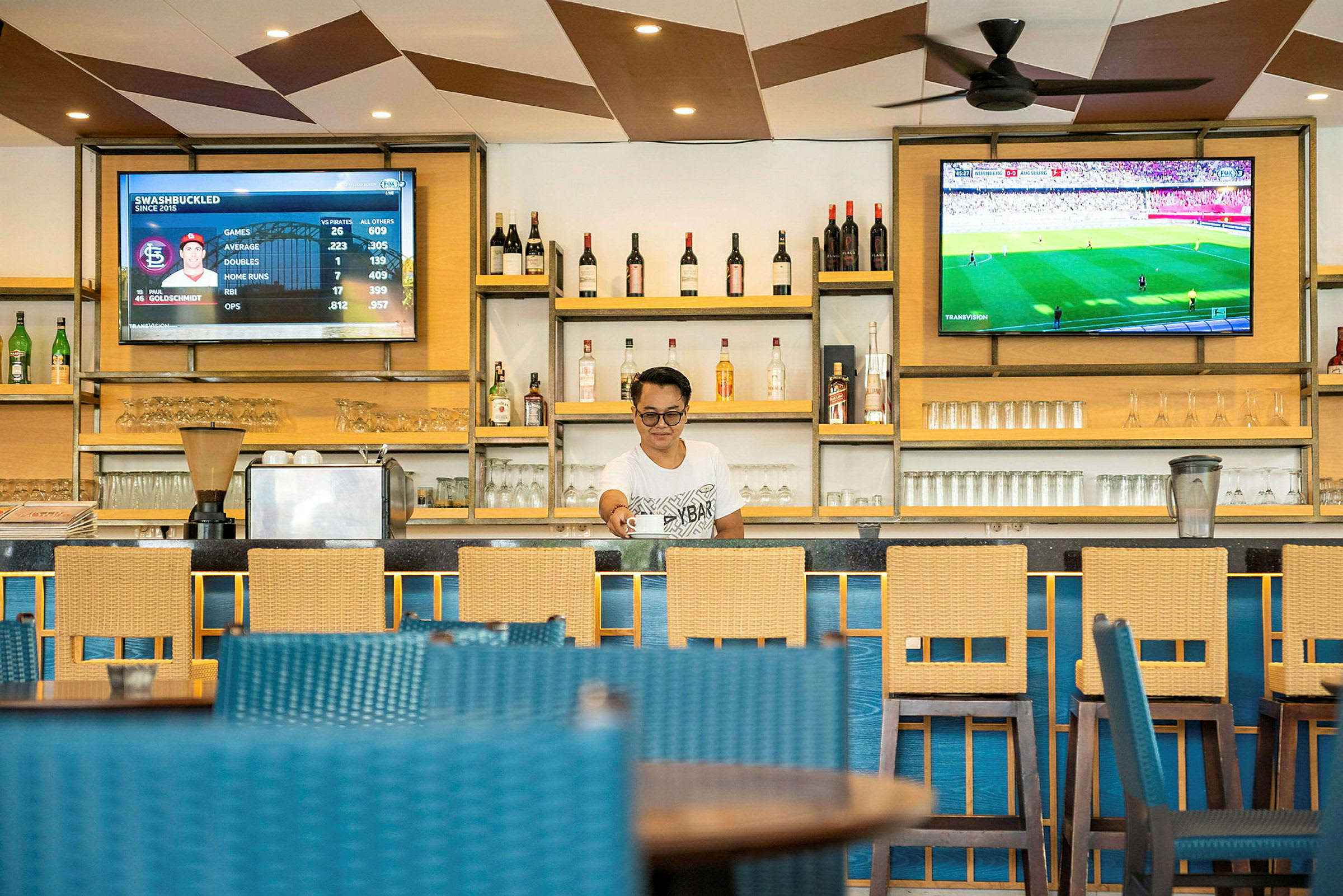 En bar med elegante tremøbler, flasker og glass på hyller, samt to skjermer som viser sportsdata og en live kamp. (AI-generert tekst)