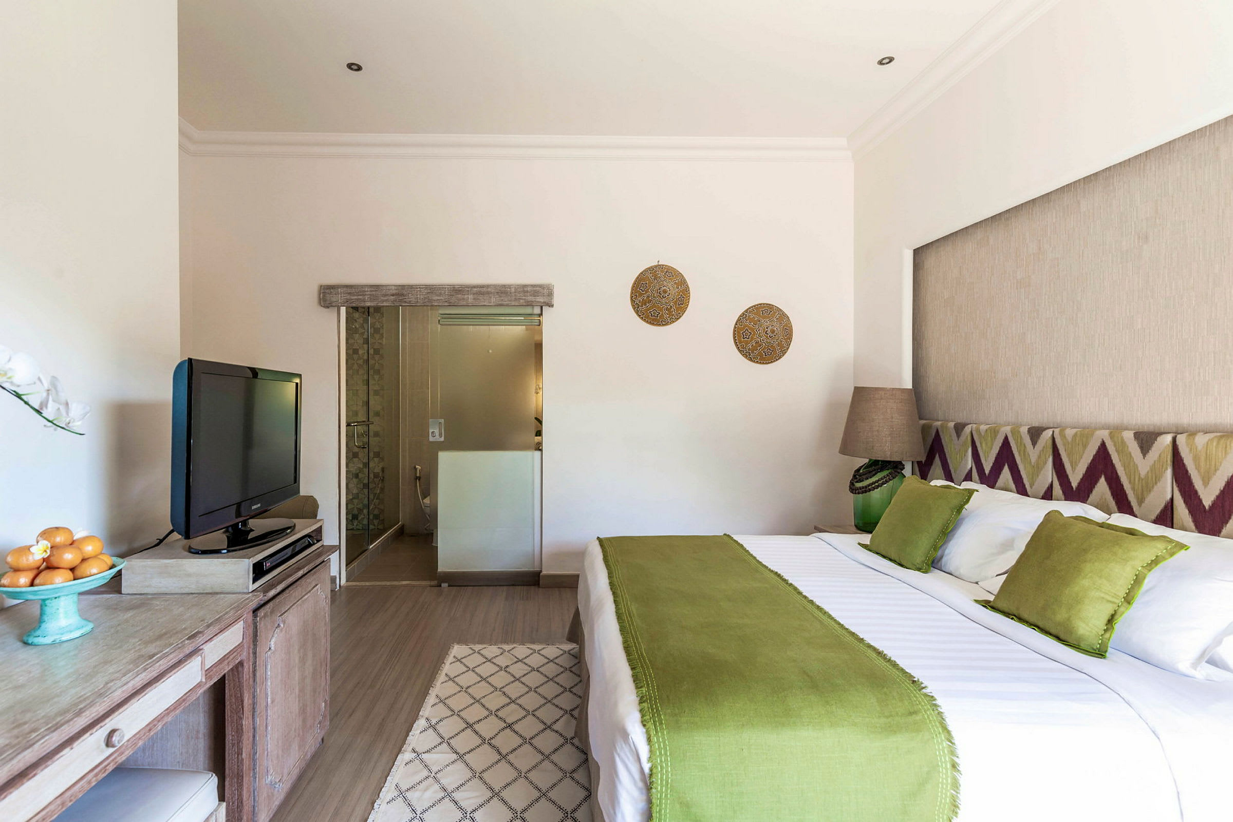 Et lyst innredet hotellrom med en stor seng, grønt sengeteppe og dekorative puter. Et skrivebord med en TV er synlig, og i bakgrunnen er det et baderom. (AI-generert tekst)