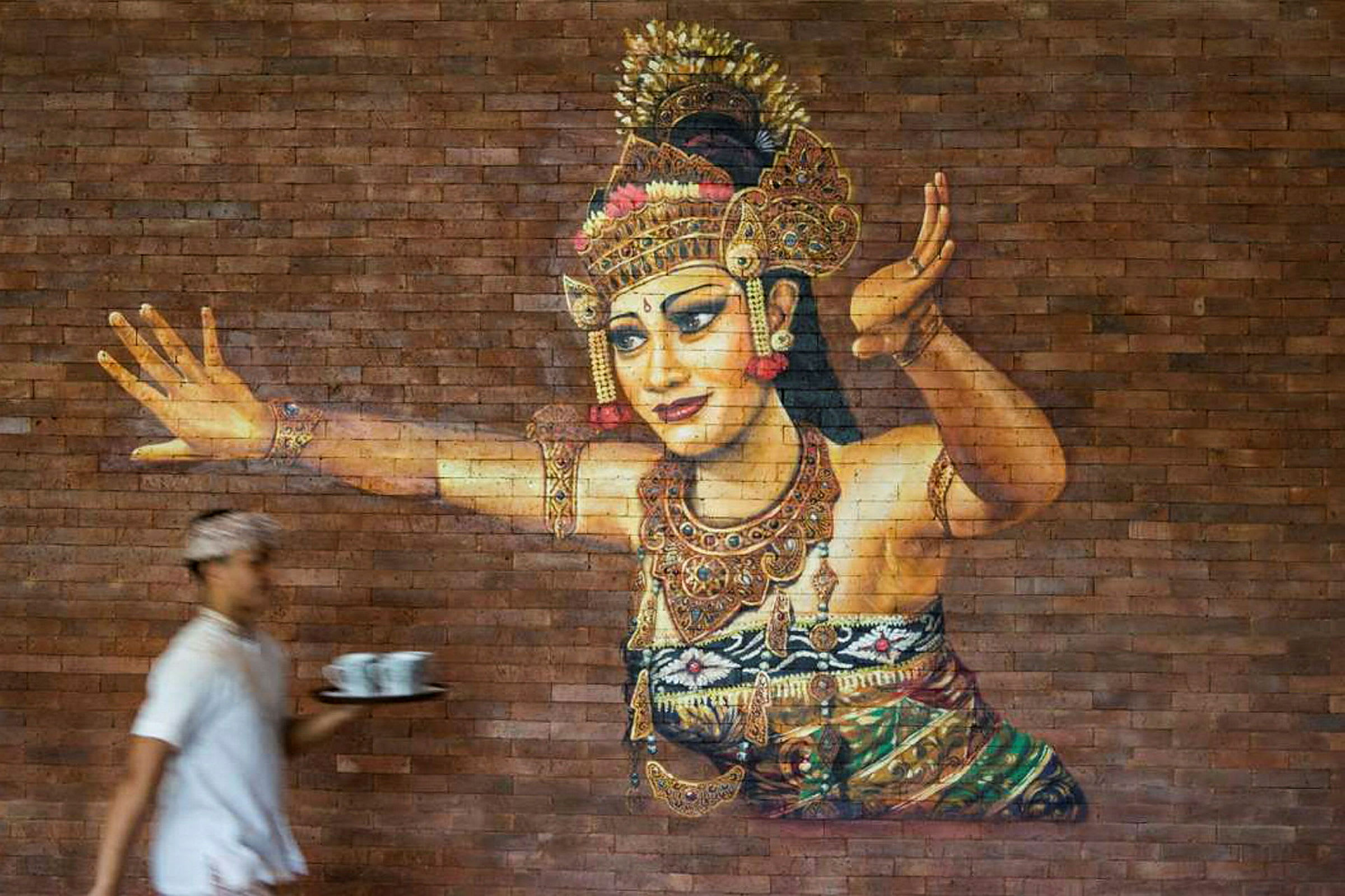 Et kunstverk på en murvegg som viser en tradisjonell danser i pyntede klær og smykker. En person i forgrunnen holder et brett med kopper. (AI-generert tekst)