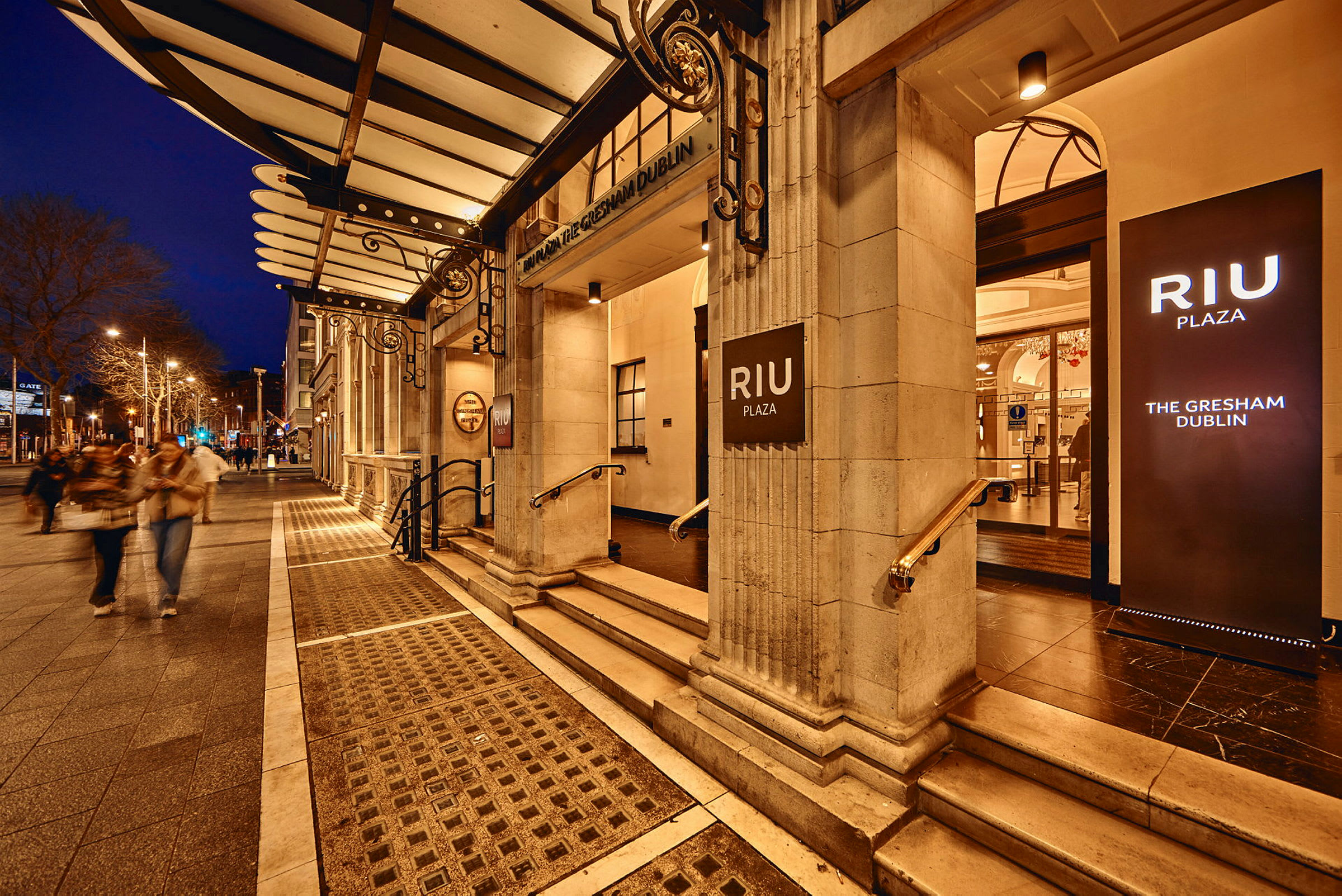 Hotel RIU Plaza Gresham Dublin inngang med opplyste skilt, kveldsmiljø. (AI-generert tekst)
