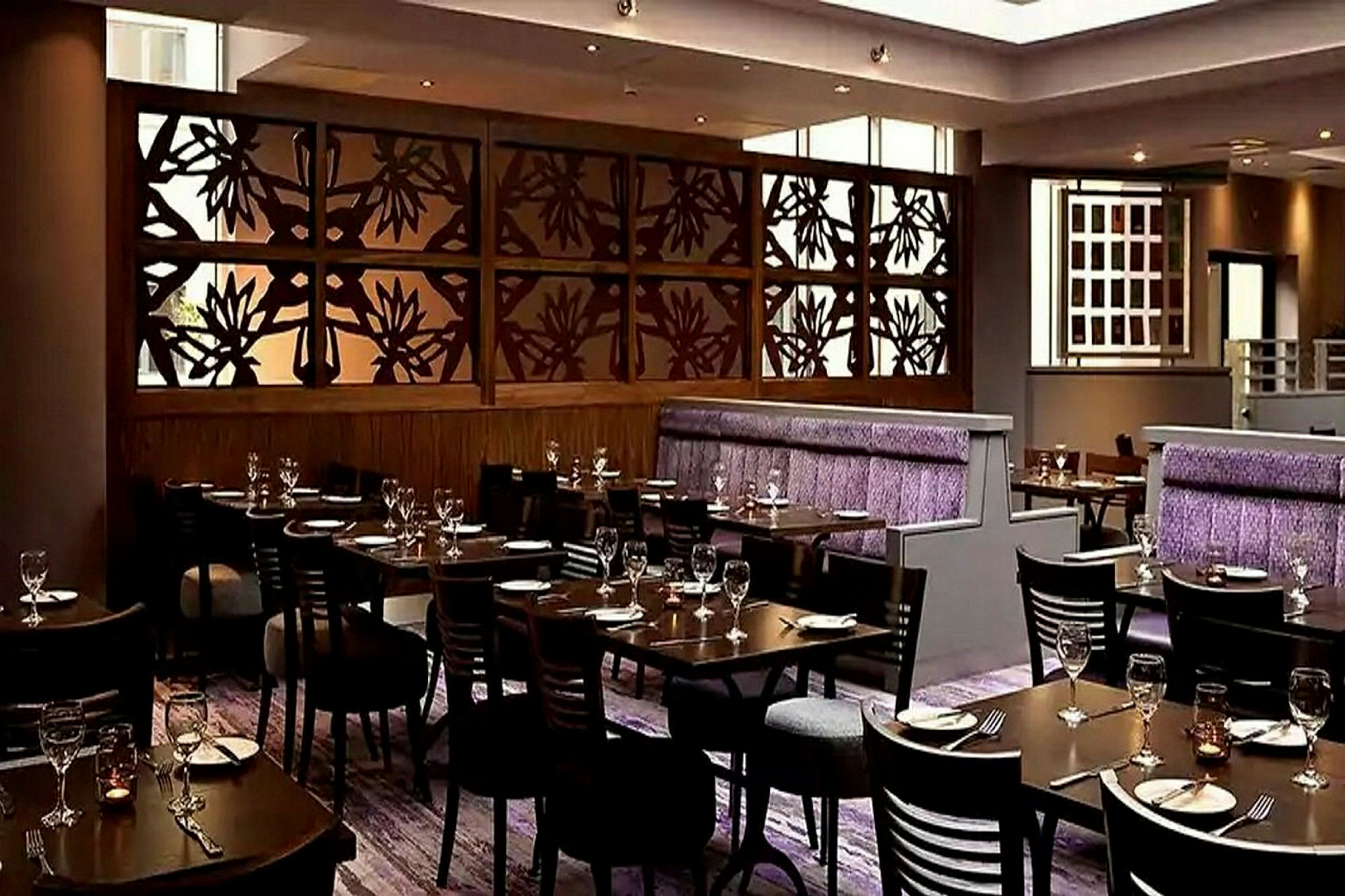 En stilfull restaurant med bord og stoler oppstilt, en dekorativ trevegg med blomstermønster og lilla detaljer på sitteplasser. (AI-generert tekst)