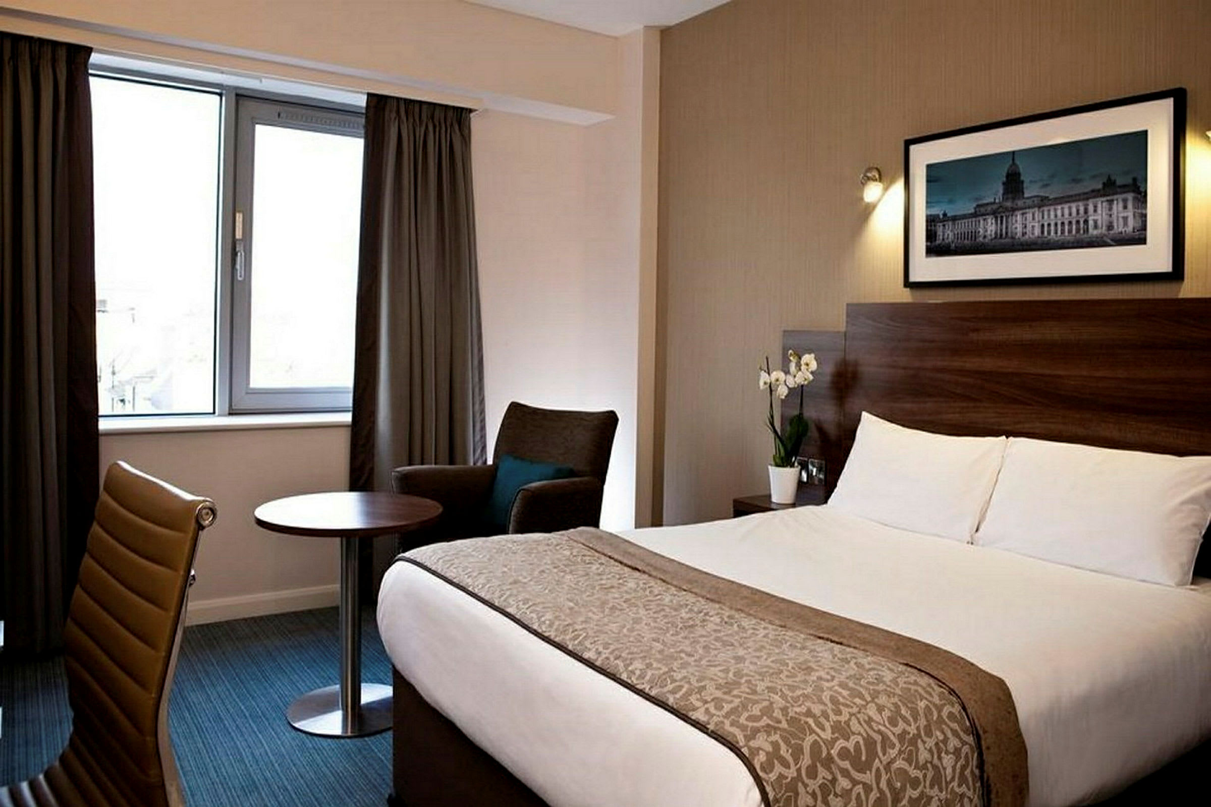 Hotellrom med seng, stol, bord og vindu med gardiner. (AI-generert tekst)