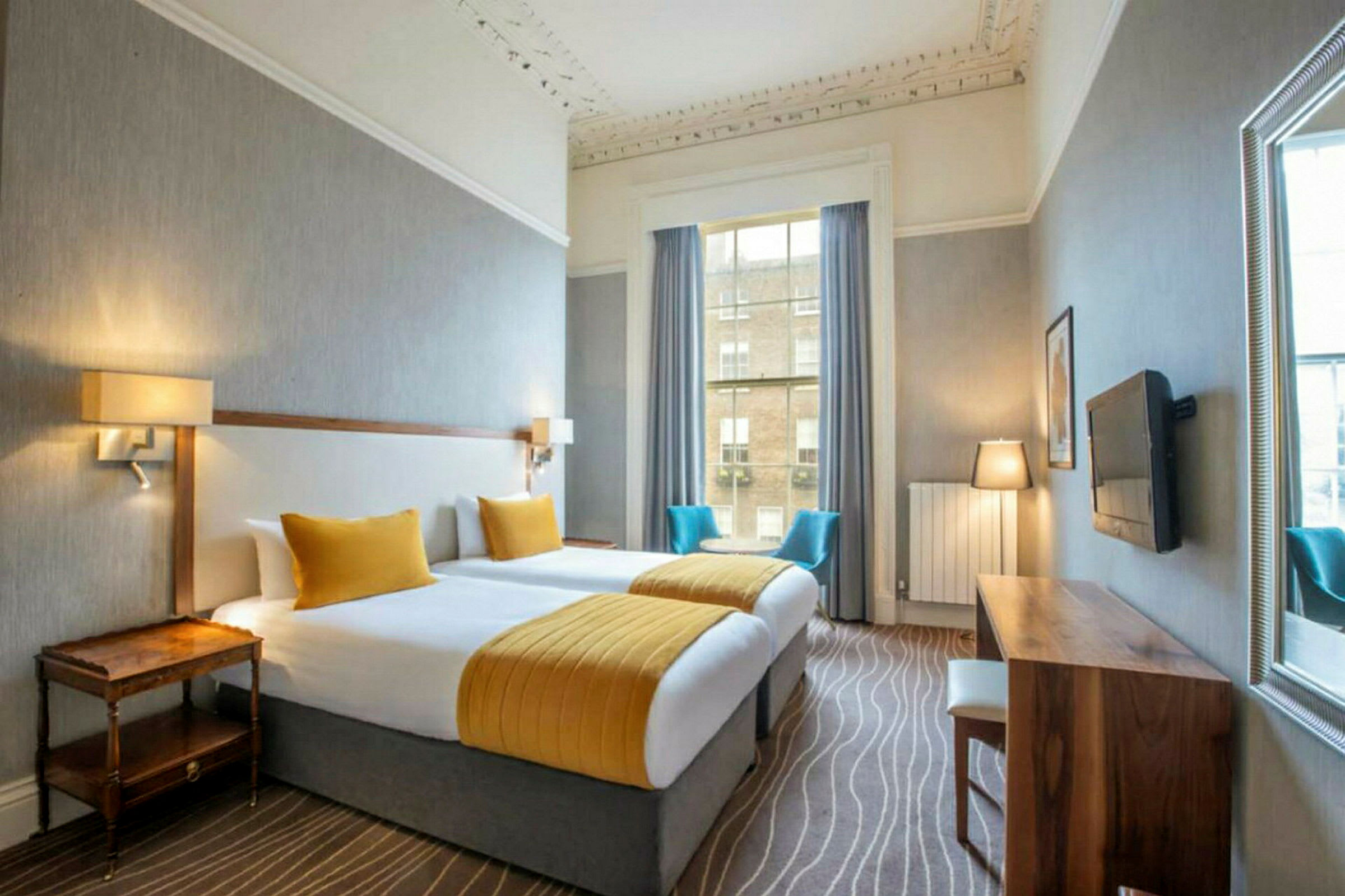 Et hotellrom med to separate senger, gullfargede sengetepper og puter, et skrivebord i tre med stol, TV på veggen, og en stor vindusåpning med blå gardiner og utsikt utover. (AI-generert tekst)