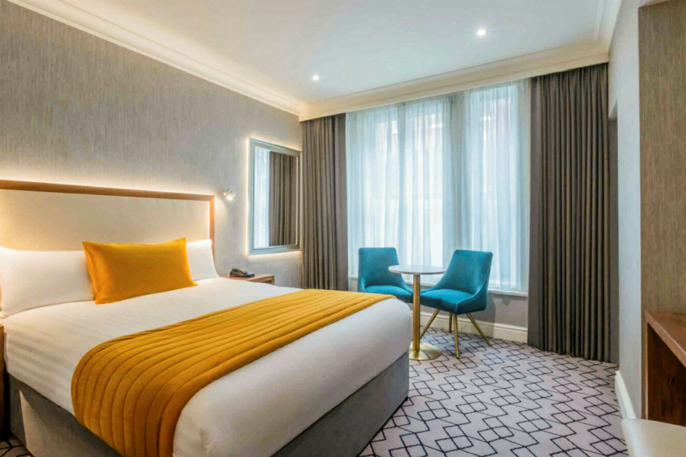 Hotellrom med en dobbeltseng, to blå stoler og et lite bord ved vinduet. (AI-generert tekst)