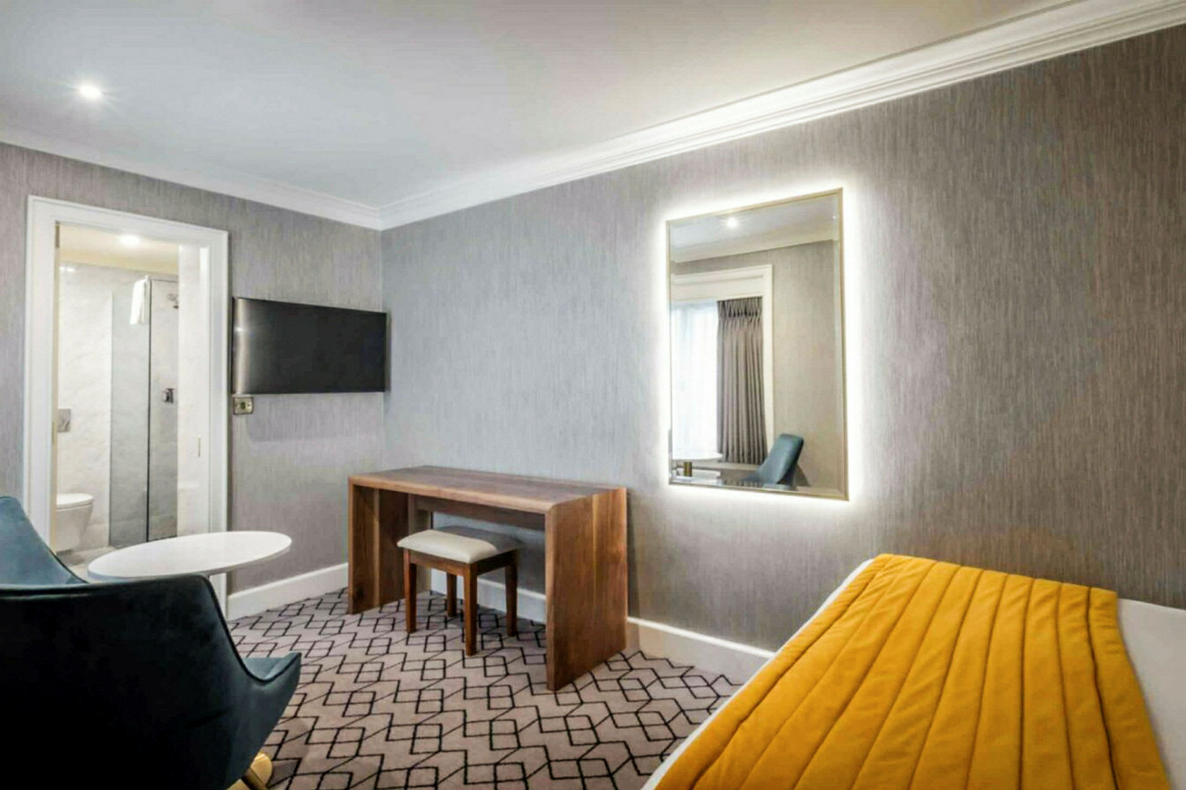 Et hotellrom med grå vegger, et speil, et skrivebord, en stol og en seng med gul teppe. (AI-generert tekst)