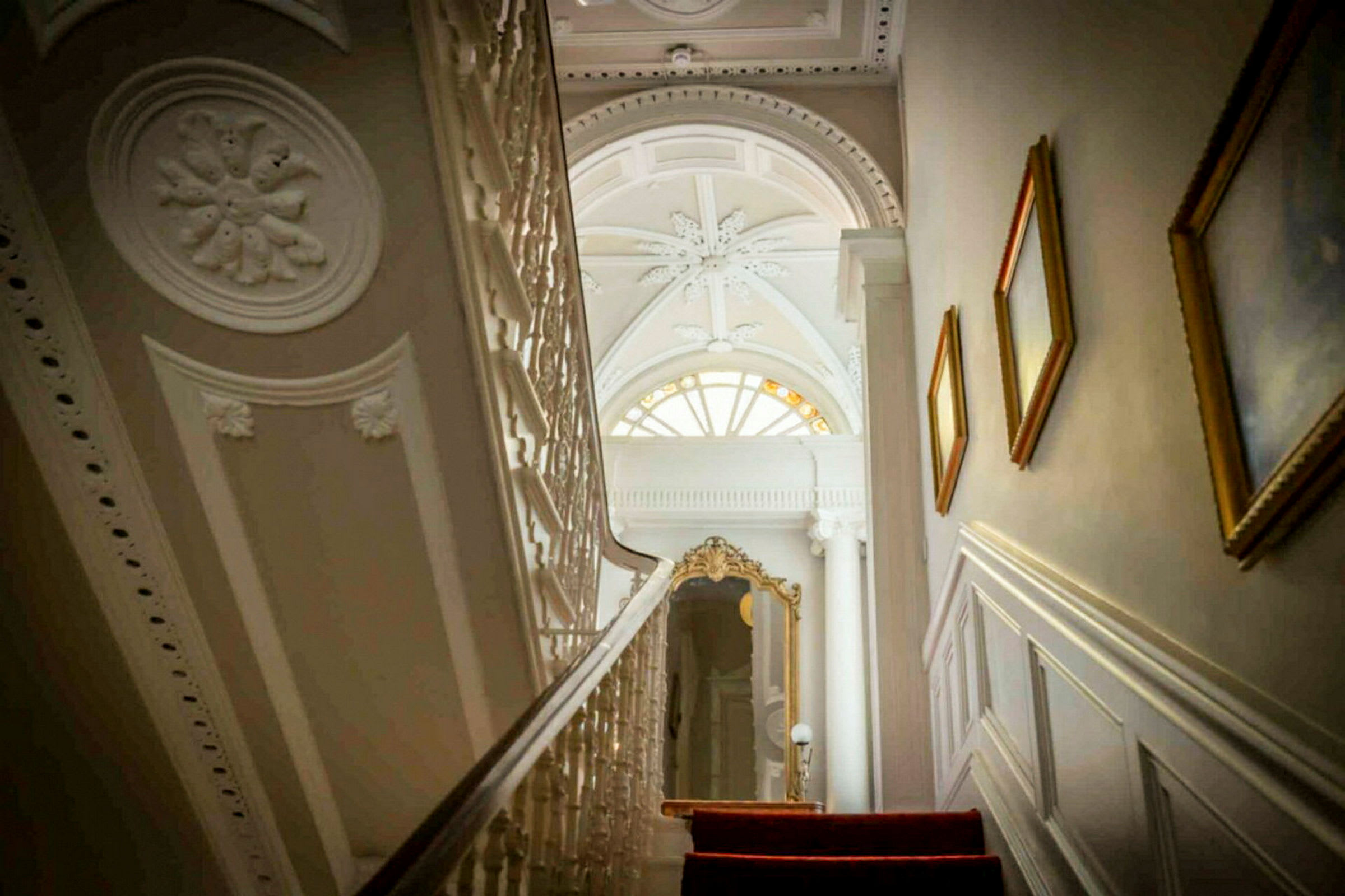 En trapp med rød teppe fører opp til et rom med elegant tak og en stor vindusformasjon. (AI-generert tekst)