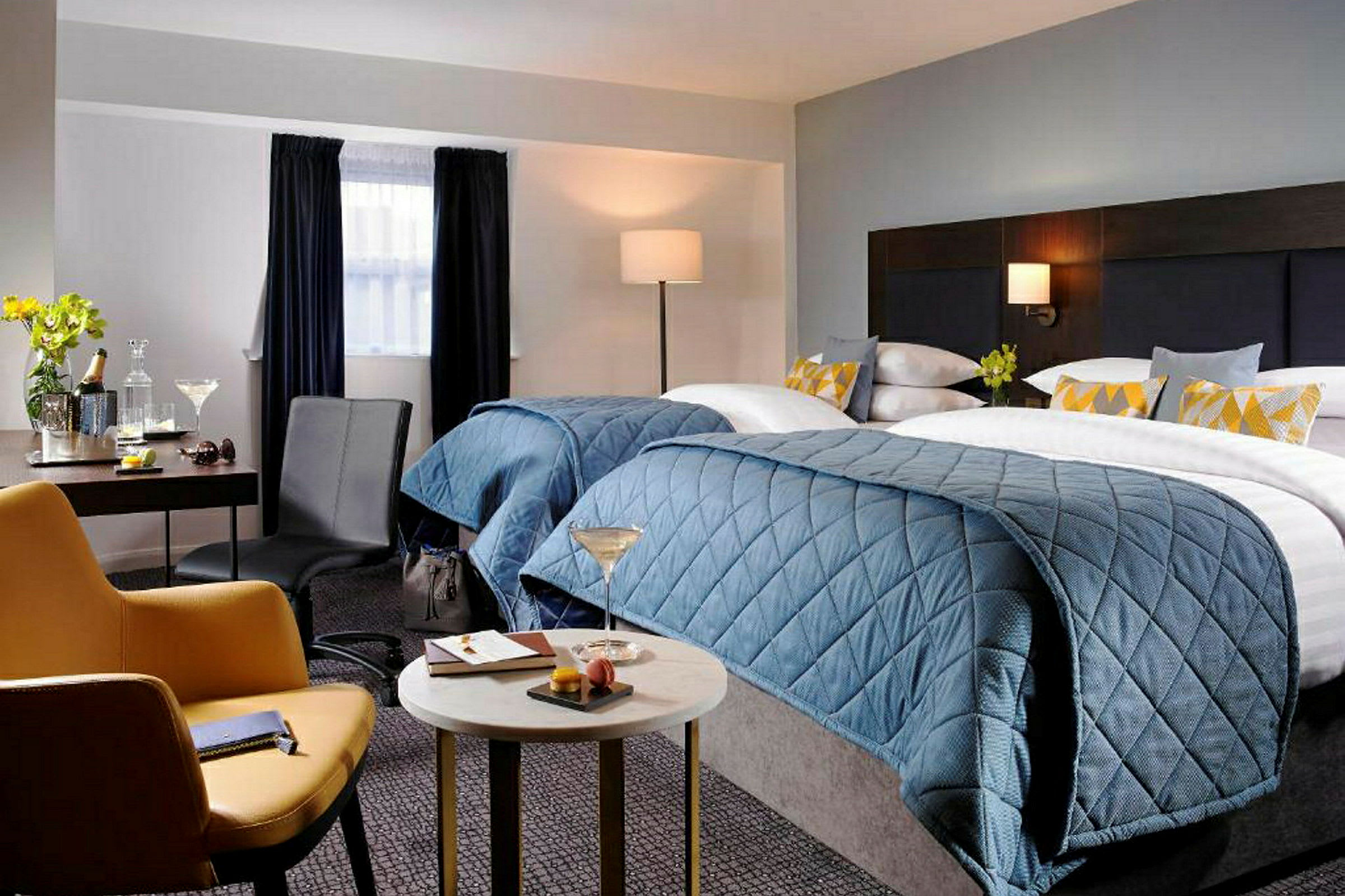 Hotellrom med to senger med blått sengeteppe, en lenestol, et bord og en arbeidsplass. (AI-generert tekst)