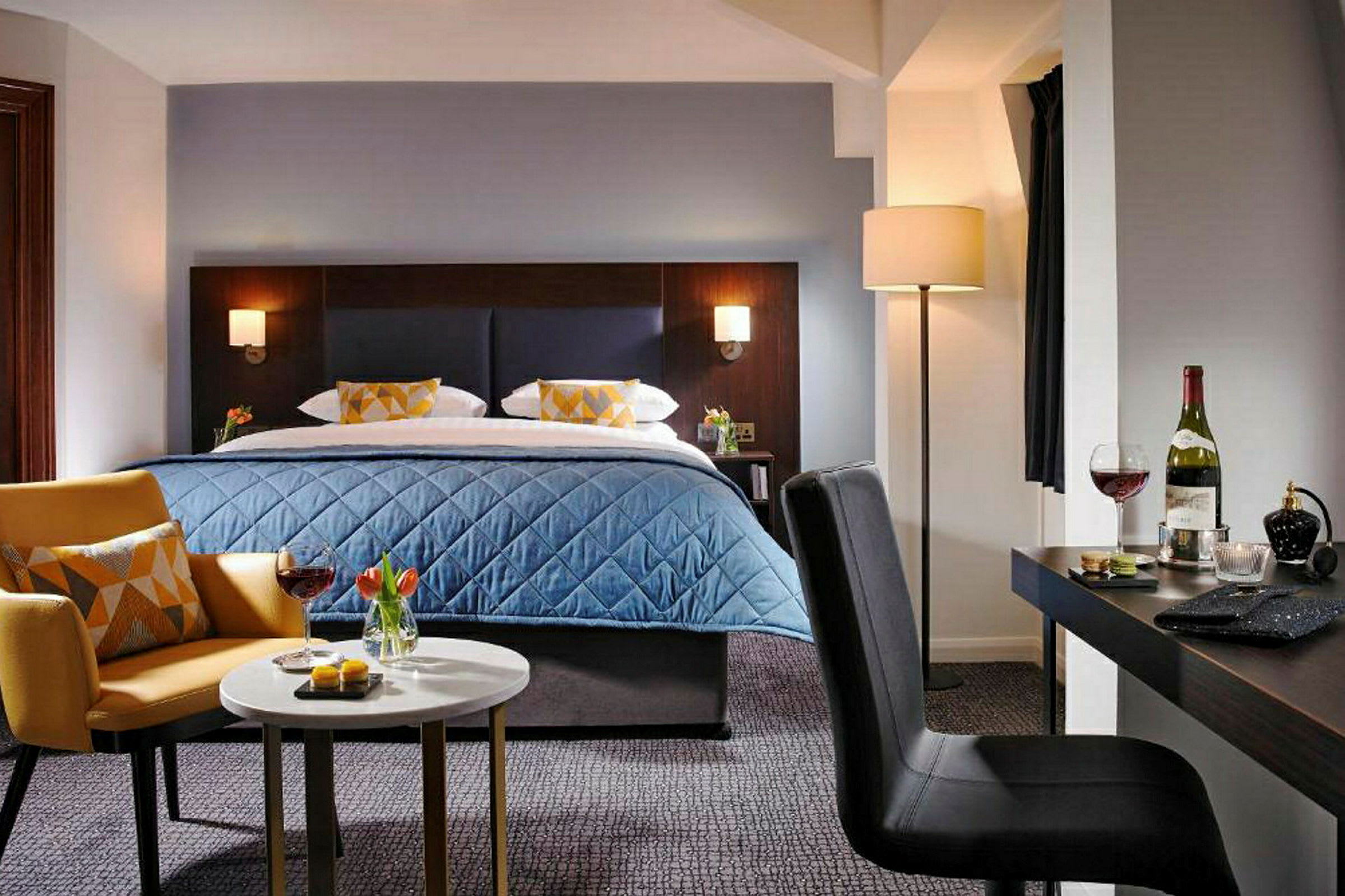 Et moderne hotellrom med en oppredd seng, et sofabord med drikke og snacks, en lenestol og et skrivebord med en stol. (AI-generert tekst)