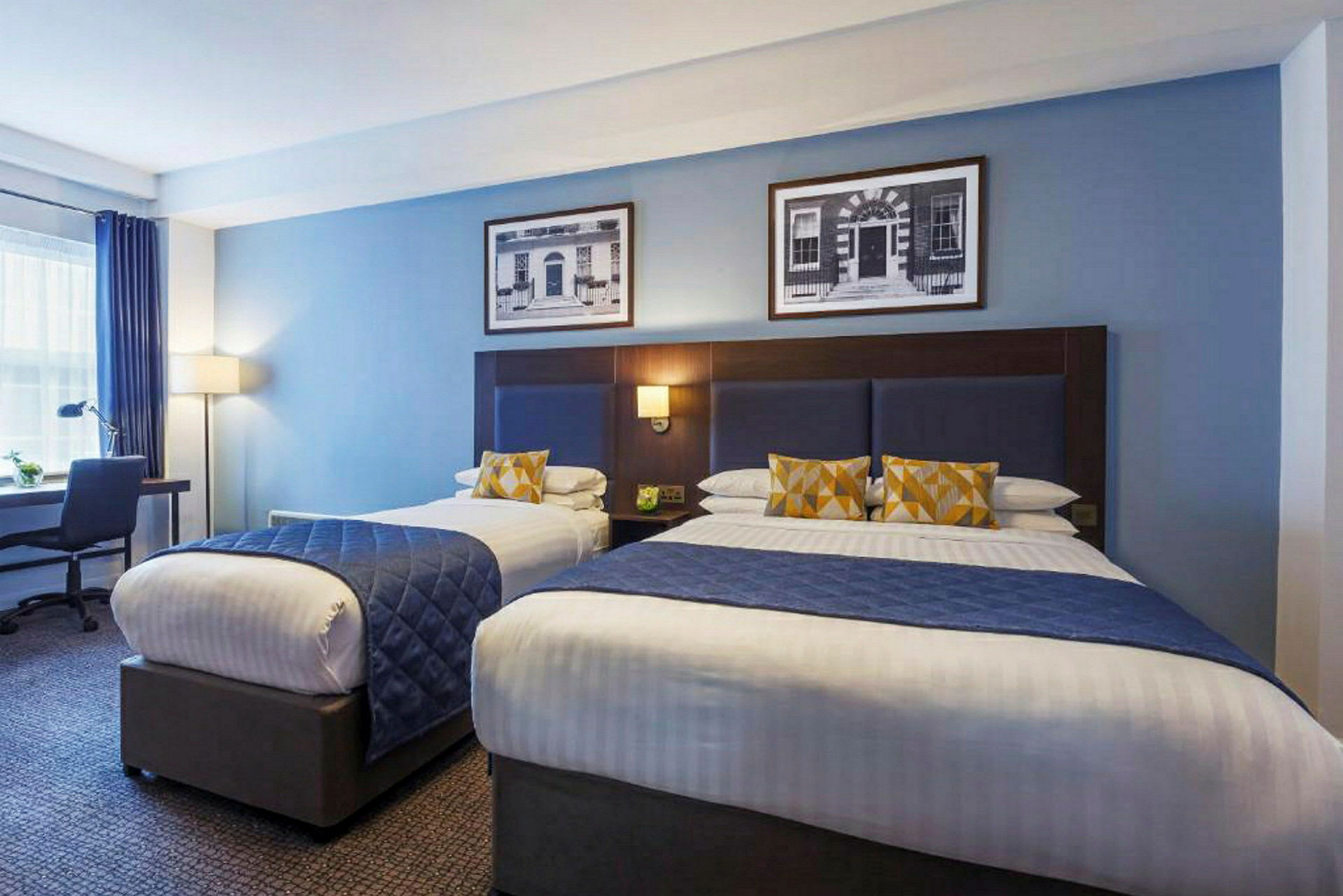 Et hotellrom med to senger, hver dekket med blå sengetepper og prydputer i gult og hvitt mønster. Ved sengen er det et skrivebord med en lampe. (AI-generert tekst)