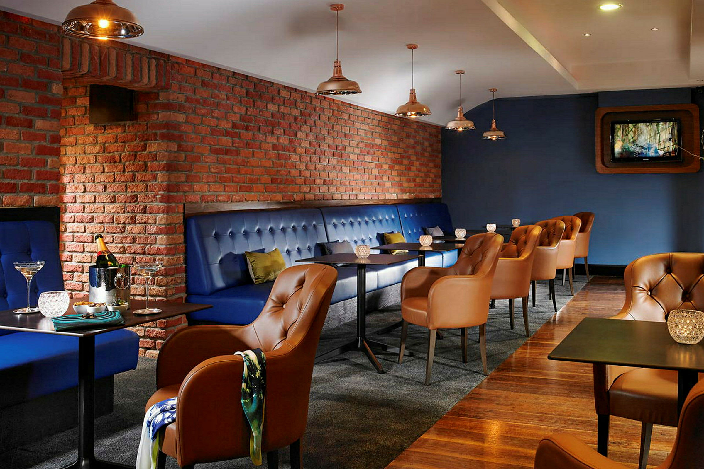 En restaurant eller lounge med blå polstrede benker og brune skinnstoler. Veggene er av rød murstein, og det er små bord med dekorative lys. (AI-generert tekst)