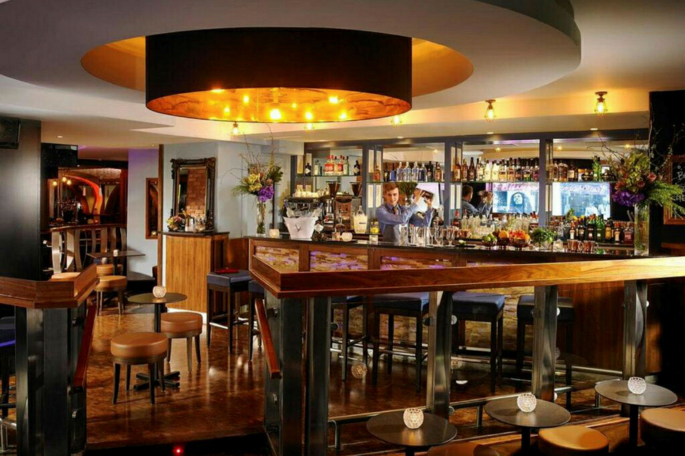 En elegant bar med treinteriør, bardisk og stoler, samt en bartender som jobber. (AI-generert tekst)