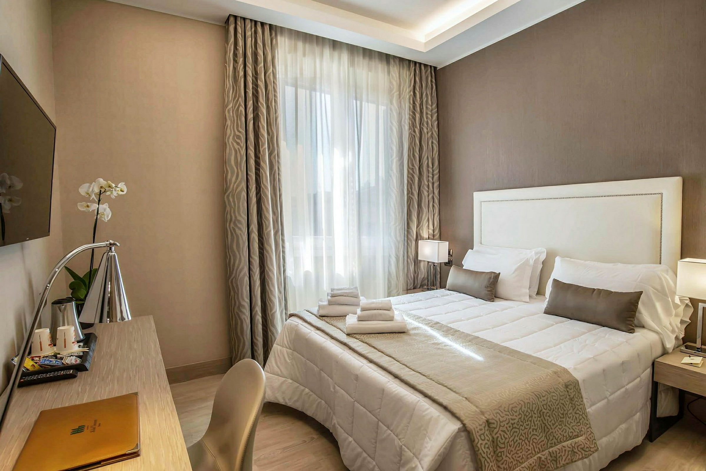 Et hotellrom med en dobbeltseng med hvitt og beige sengetøy, en skrivebord med lampe og en veggmontert TV. (AI-generert tekst)