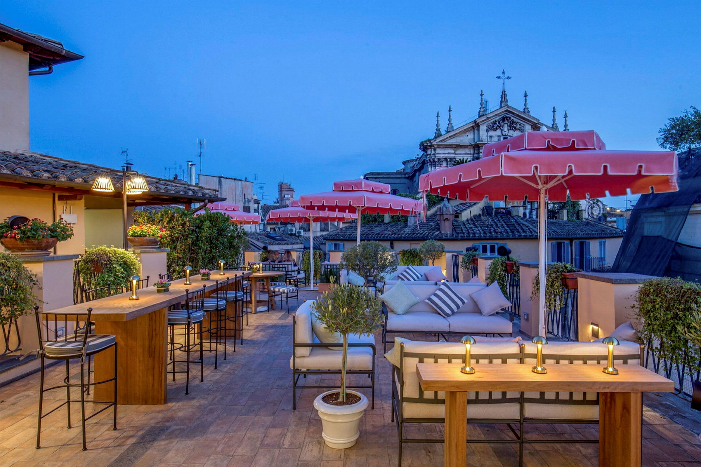 En terrasse med bord og stoler, røde parasoller, grønne planter og utsikt over byens bygninger. (AI-generert tekst)
