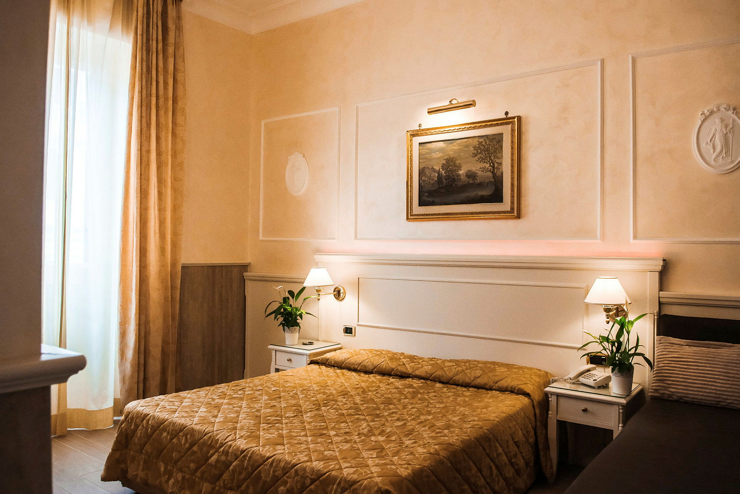 Et hotellrom med en seng dekket av et gullfarget sengeteppe, to nattbord med lamper og grønne planter, samt et maleri på veggen over sengen. (AI-generert tekst)