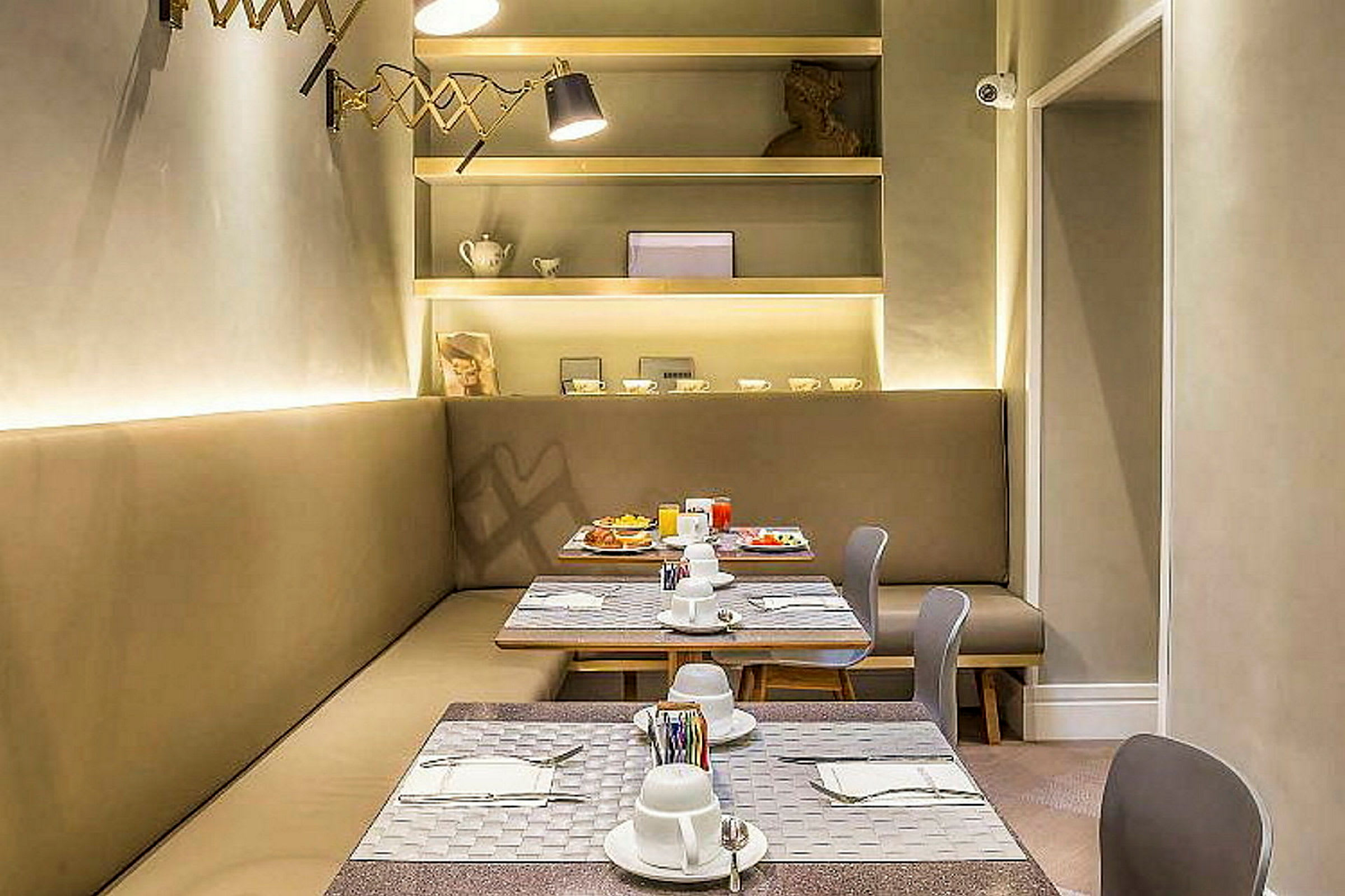 Et moderne møblert restaurantområde med bord, stoler og en sittebenk. Bordet er dekket med porselen og bestikk. På veggen er det hyller med dekorative gjenstander. (AI-generert tekst)