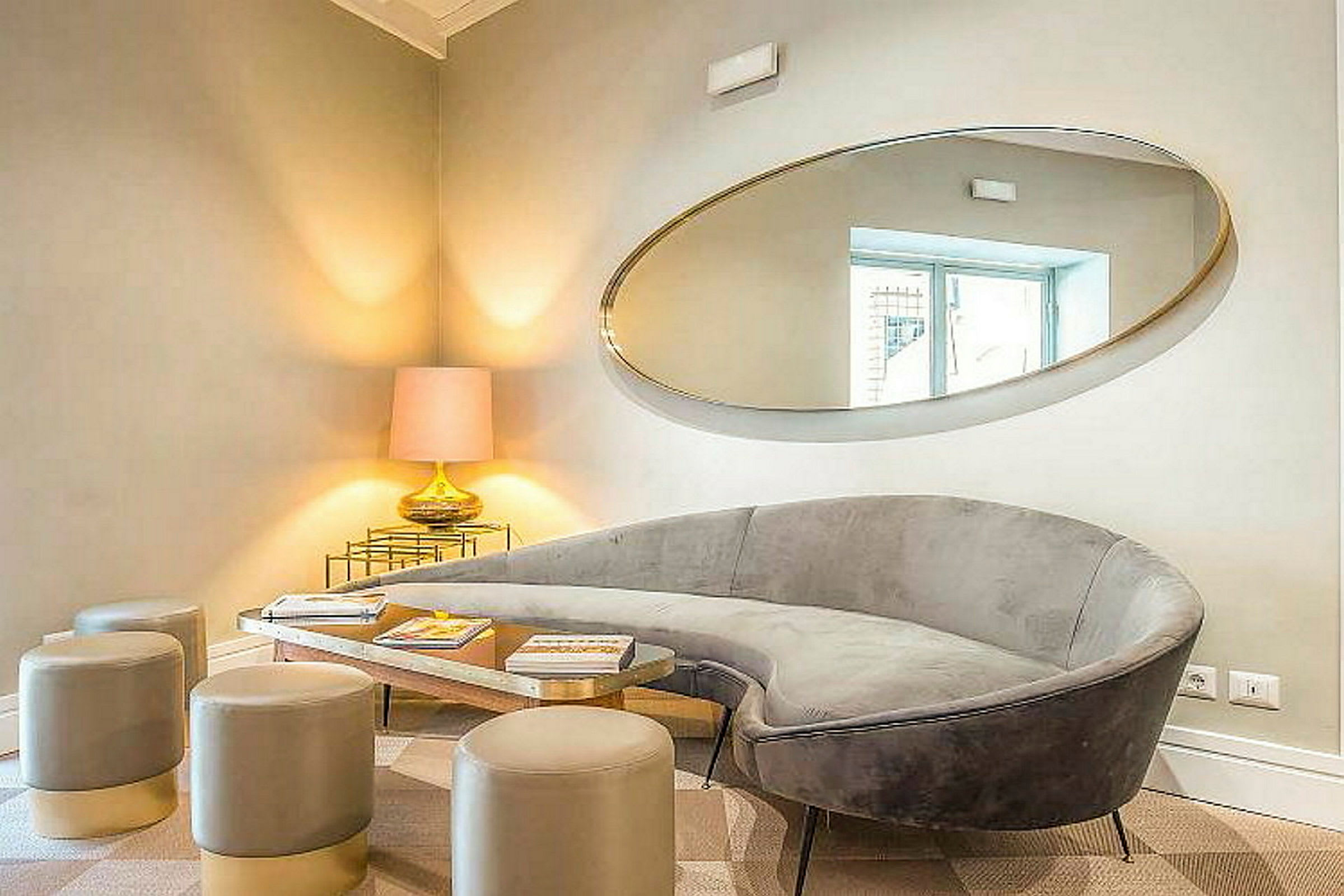 Et elegant oppholdsrom med en grå sofa, moderne sittepaller, et speilet bord og en bordlampe plassert i hjørnet. (AI-generert tekst)