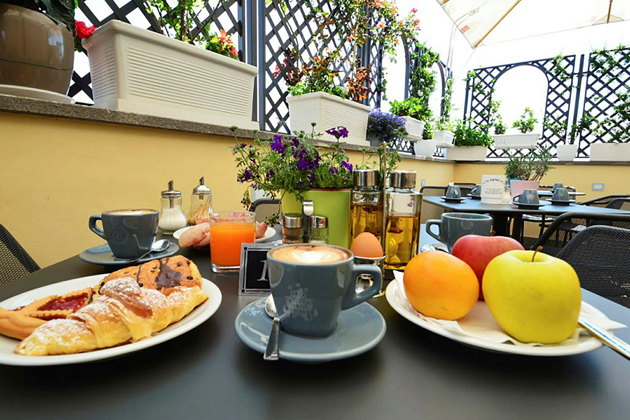 Et bord på en solrik uteplass med en frokost bestående av kaffe, juice, croissanter, frukt og andre tilbehør. (AI-generert tekst)