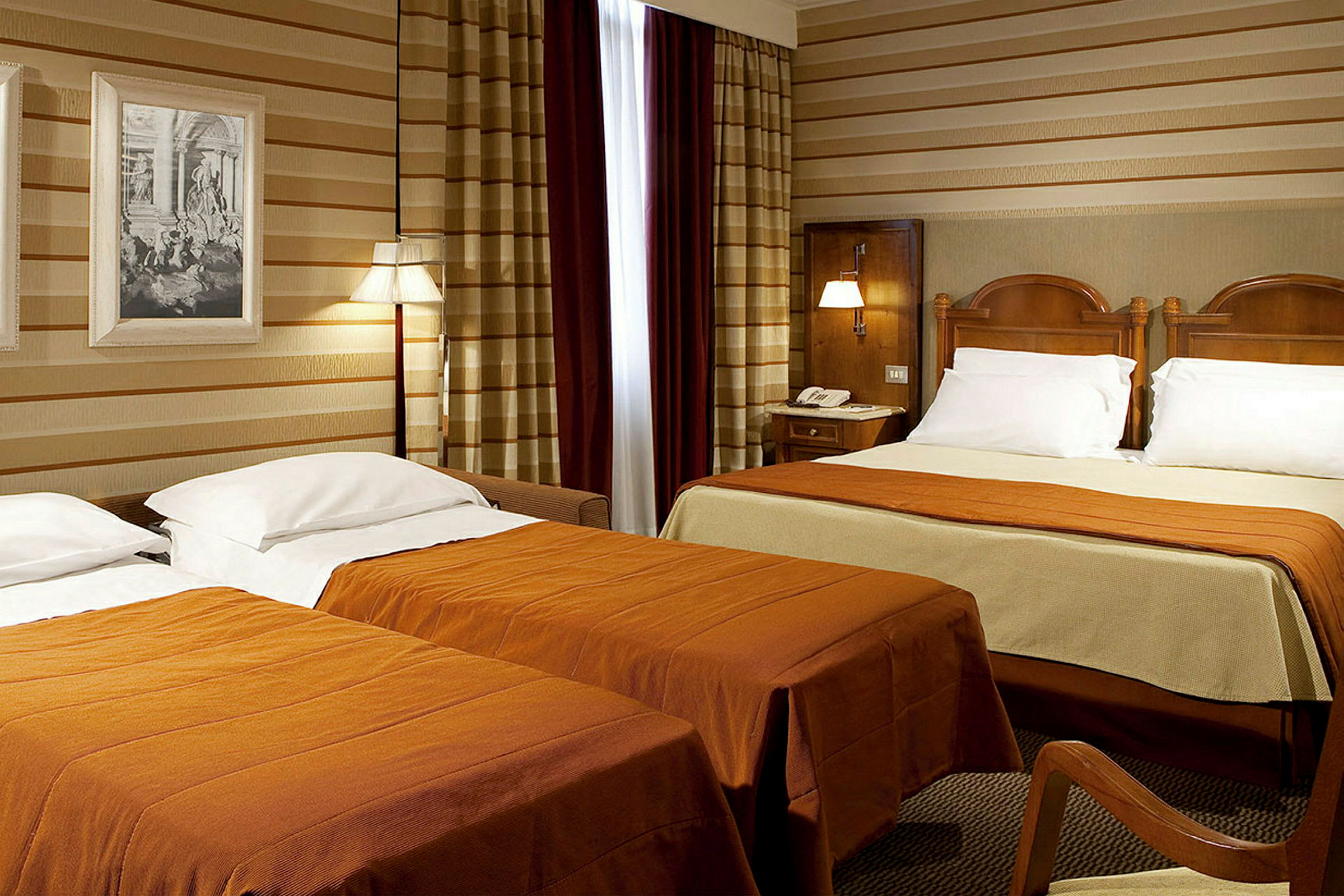 Et hotellrom med to senger, hvorav den ene er en dobbeltseng. Sengene har oransje sengetepper og hvite puter. (AI-generert tekst)