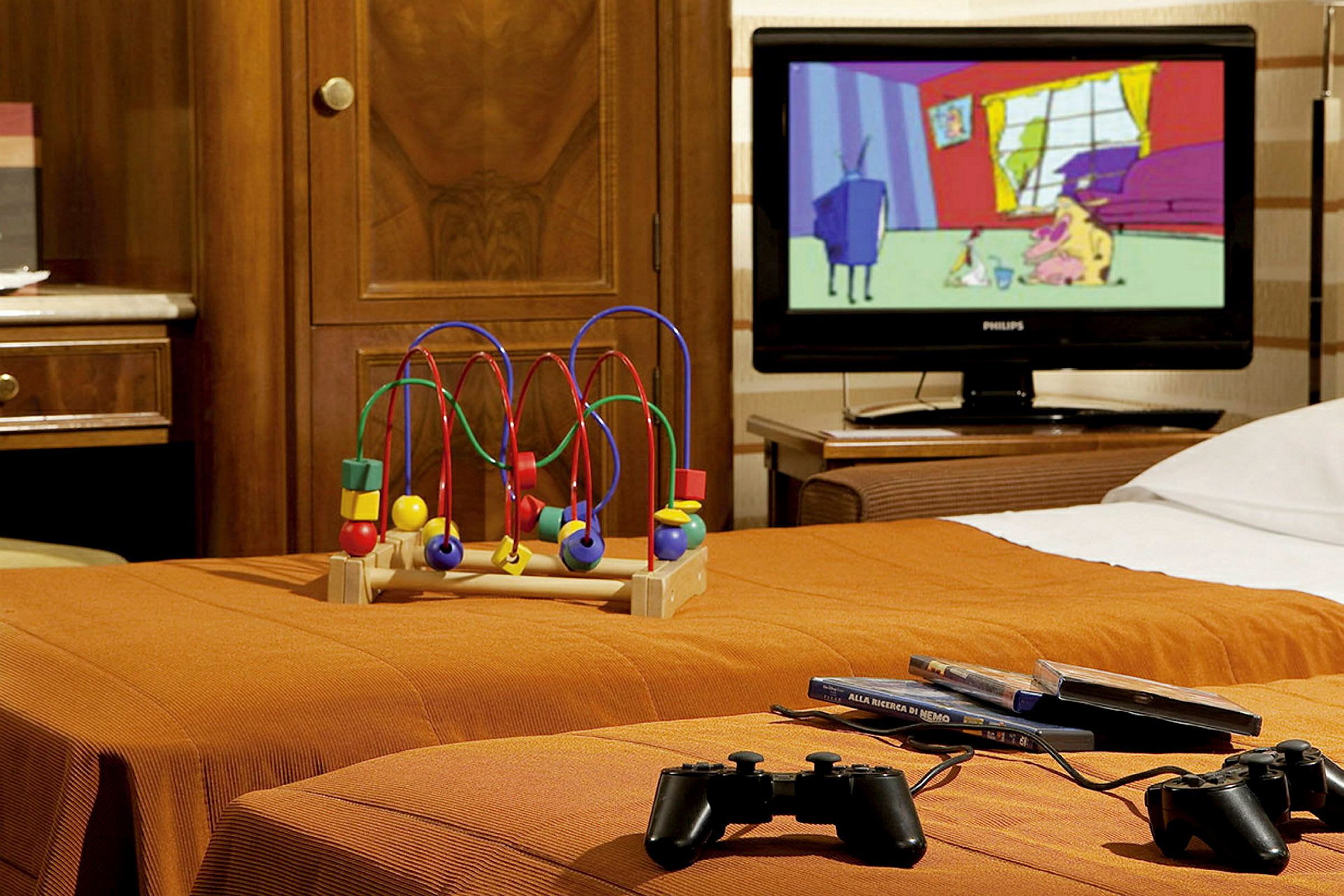 Et hotellrom med leke, TV og spillkontrollere på en orange seng. (AI-generert tekst)