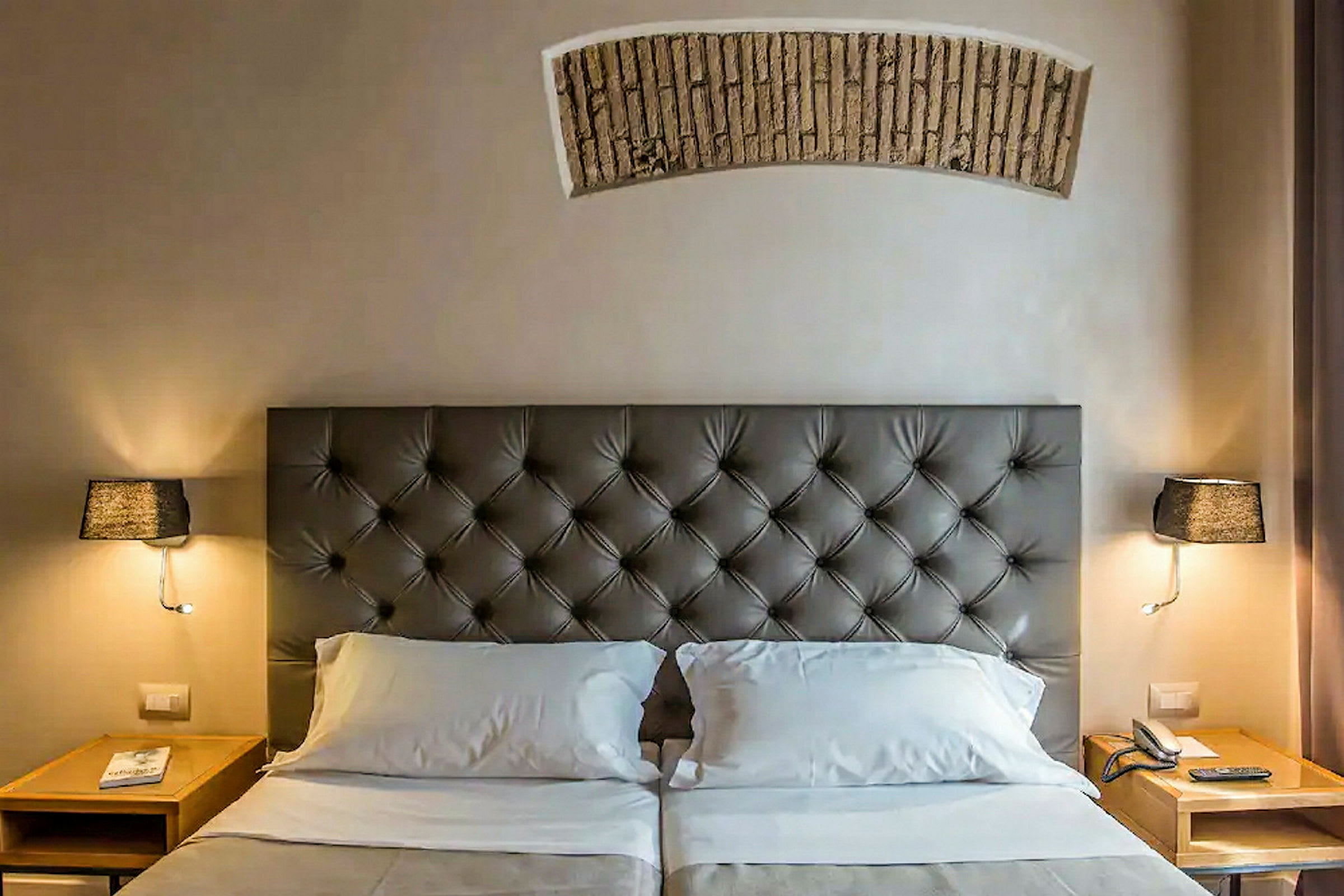 Et hotellrom med en dobbeltseng som har en polstret sengegavl, to nattbord med lamper og en telefon. (AI-generert tekst)