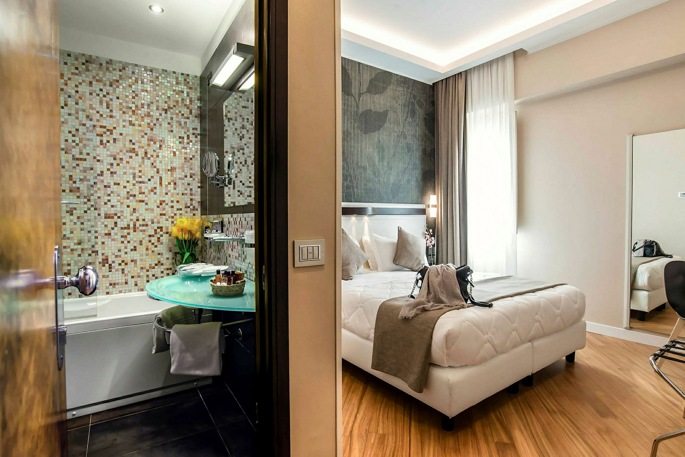 Et hotellrom med en seng foran et stort vindu, samt et bad med mosaikkvegg og en moderne vask. (AI-generert tekst)